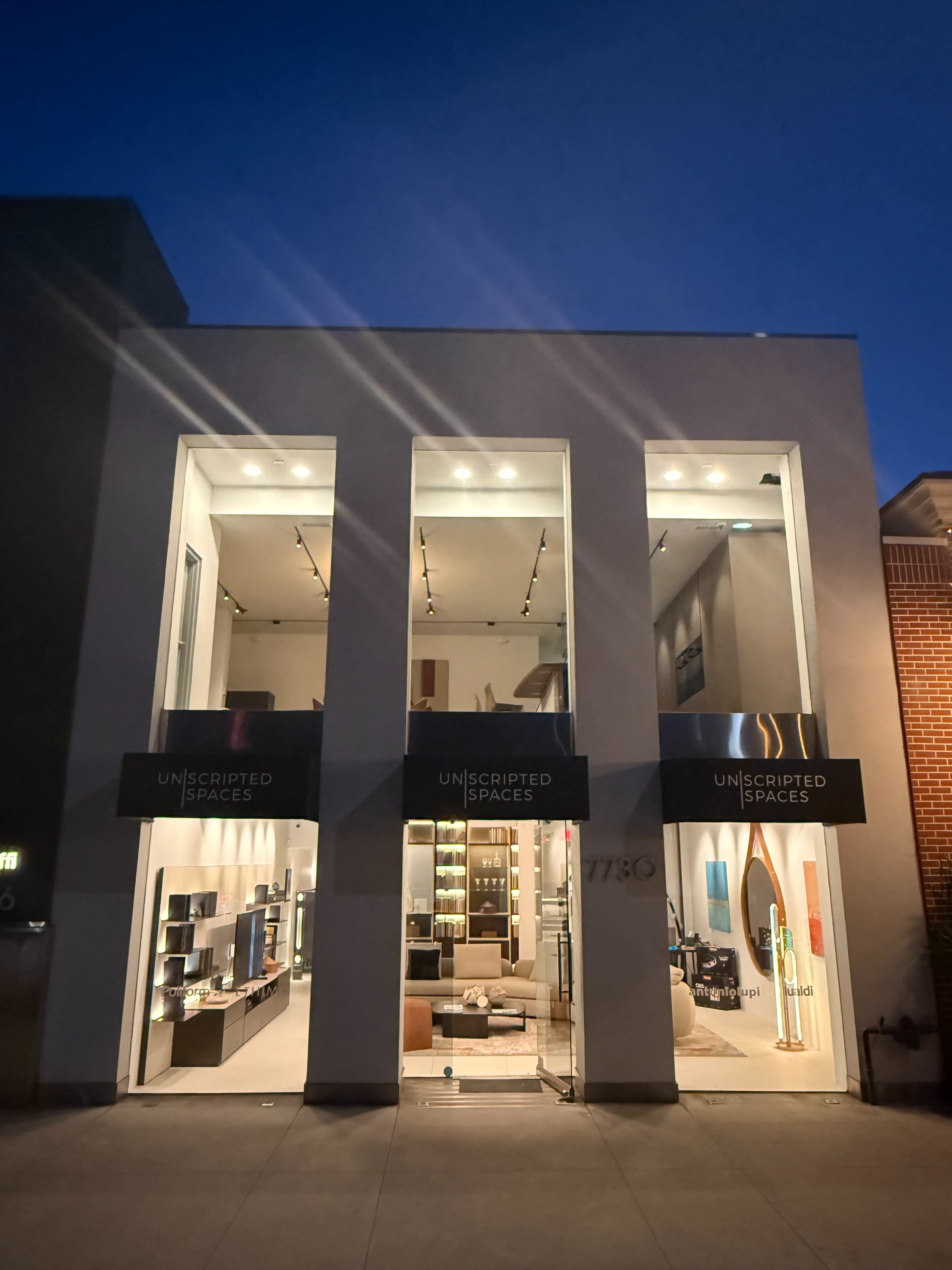 The image shows a modern storefront with large windows displaying furniture and decor, illuminated at dusk.