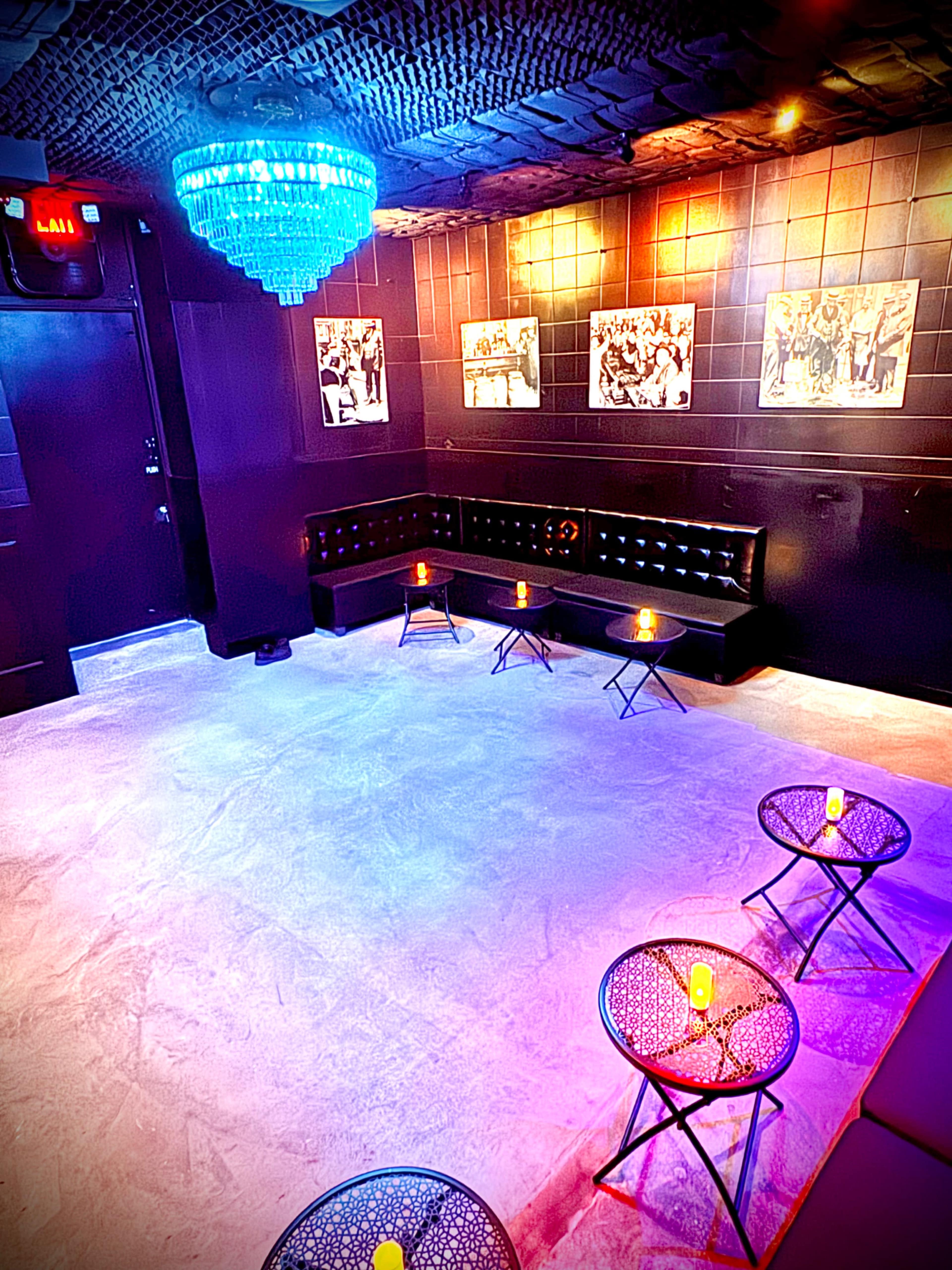 The image shows a dimly lit lounge area with a black leather couch, small round tables, and a chandelier above, decorated with wall-mounted black-and-white photographs.