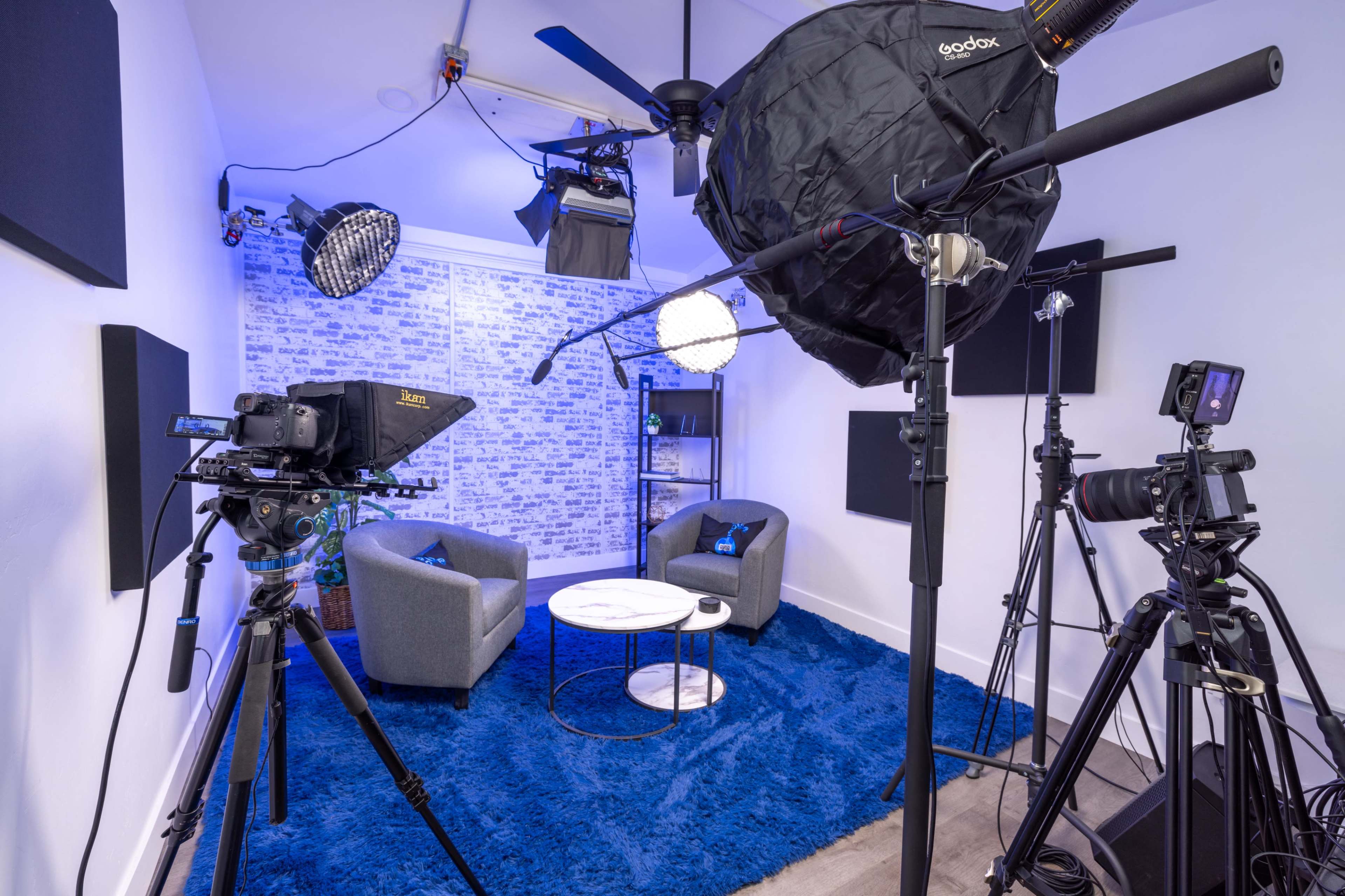 The image shows a small studio setup with two gray chairs, a round table, and various lighting and video equipment arranged around a blue carpet.