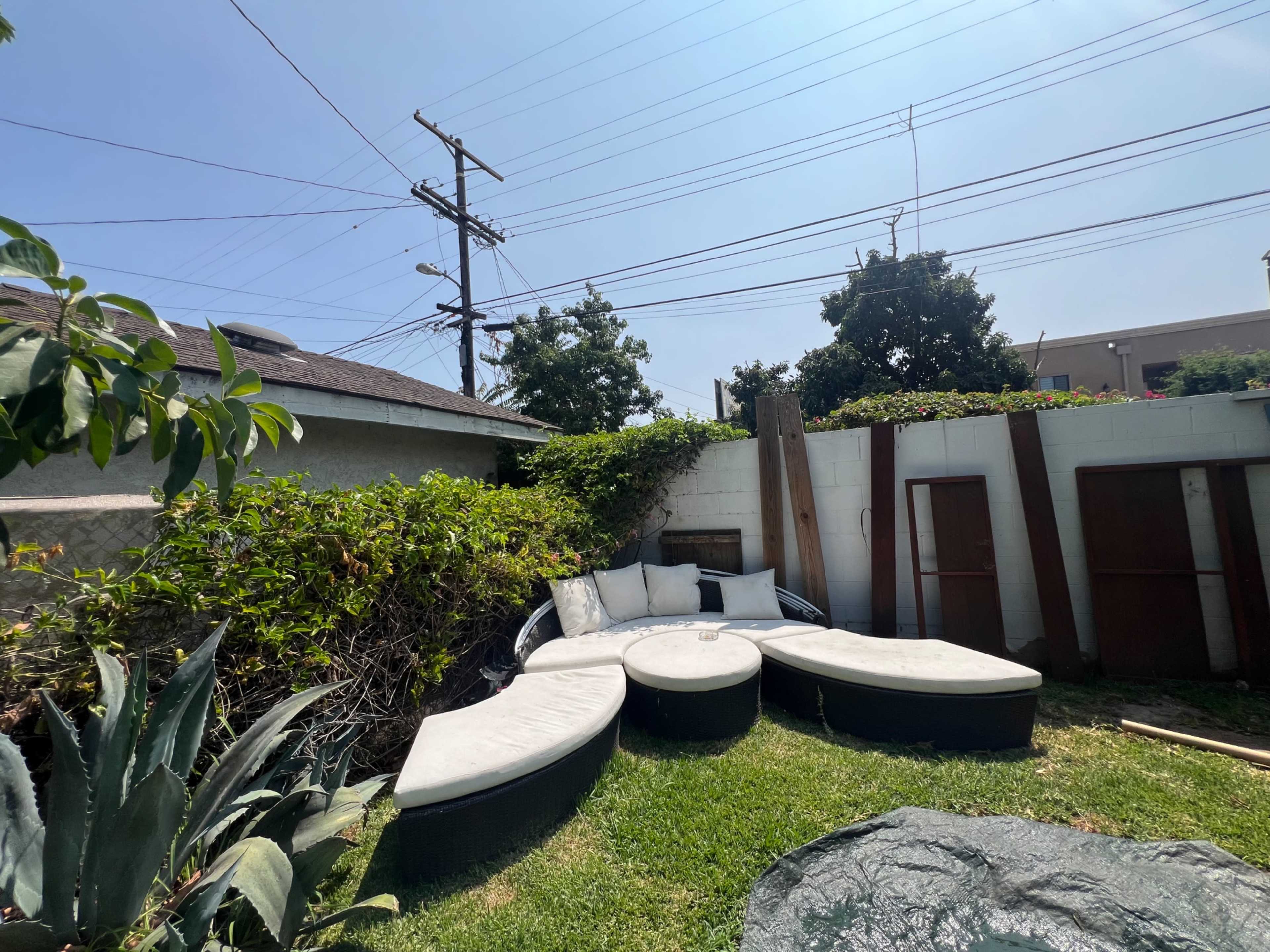 A circular outdoor seating area with black cushions is surrounded by greenery and a fence, under a clear blue sky.