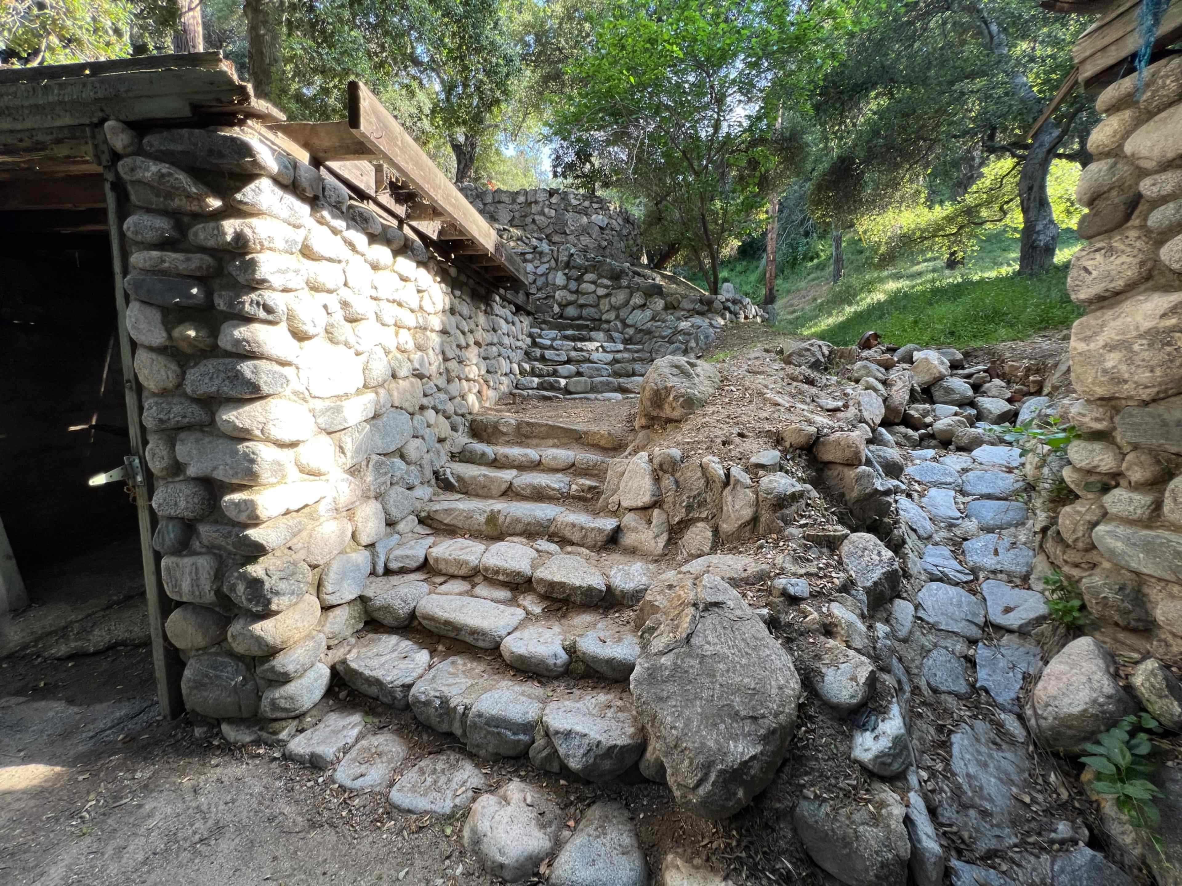 A stone pathway with steps leading up through a wooded area, flanked by stone walls and surrounding greenery.