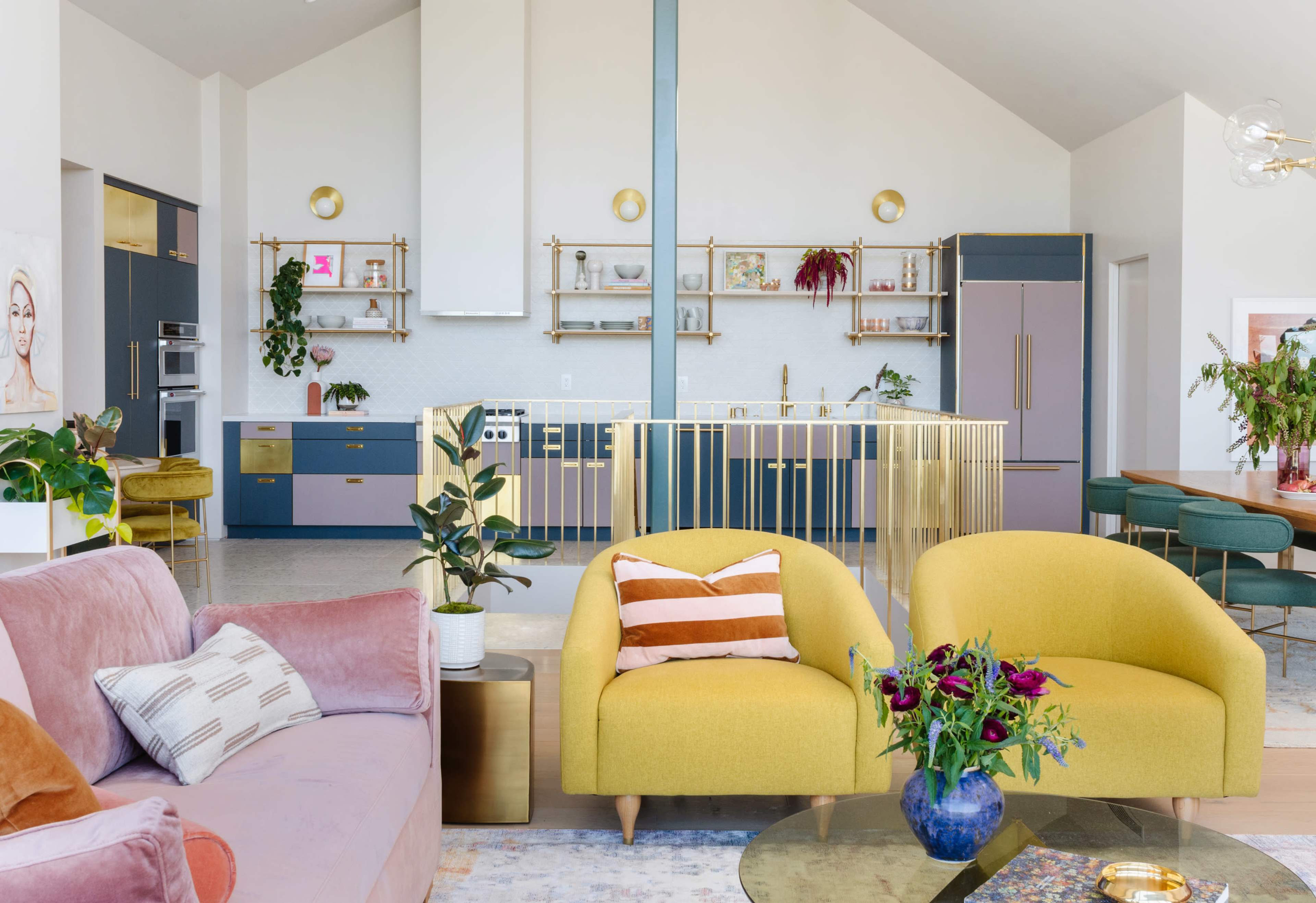 A modern living space with a kitchen and dining area, featuring yellow chairs, a pink sofa, and plants throughout.