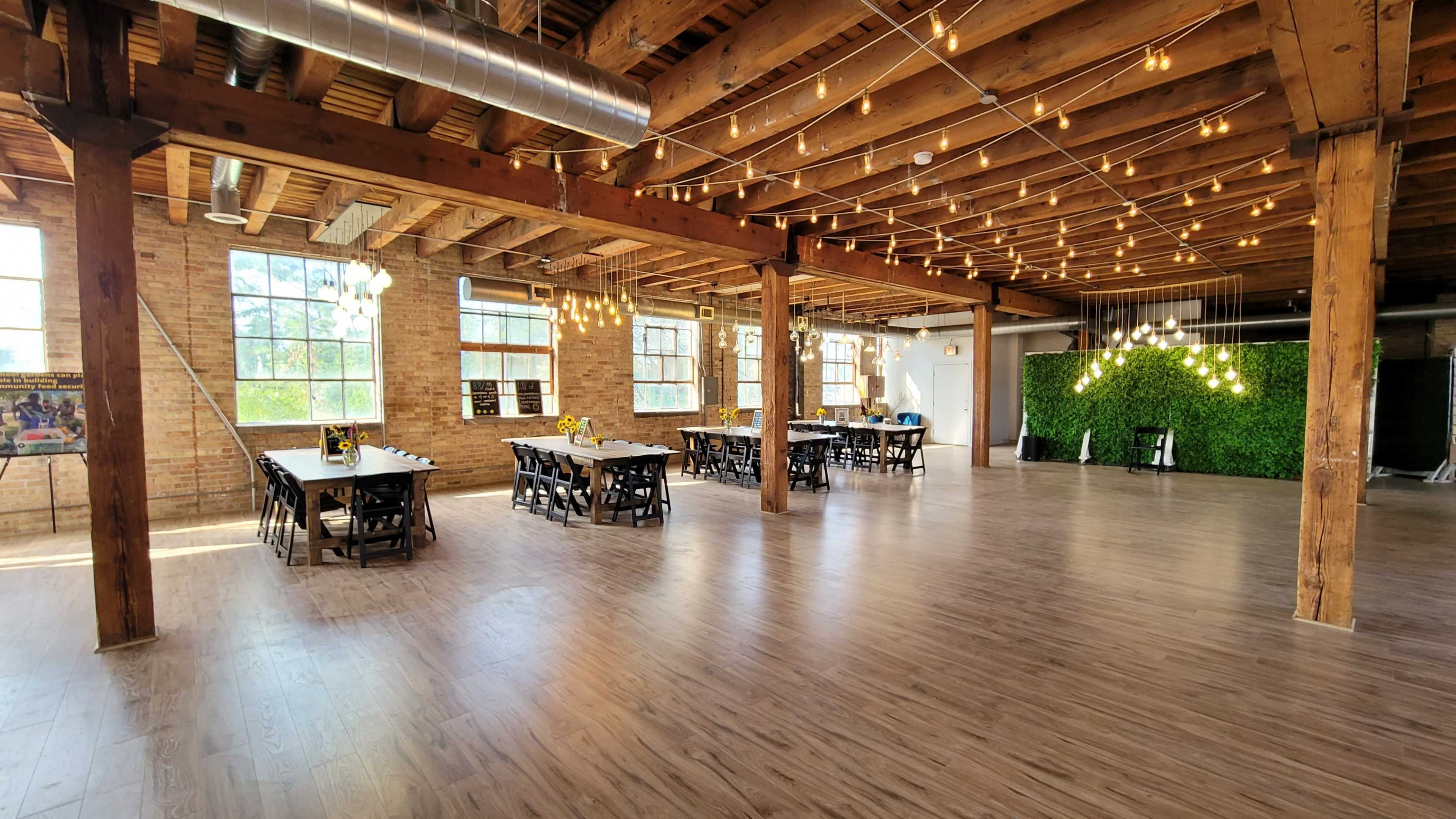 A spacious, well-lit event venue with wooden beams, industrial-style lighting, and rows of tables set up for a gathering.