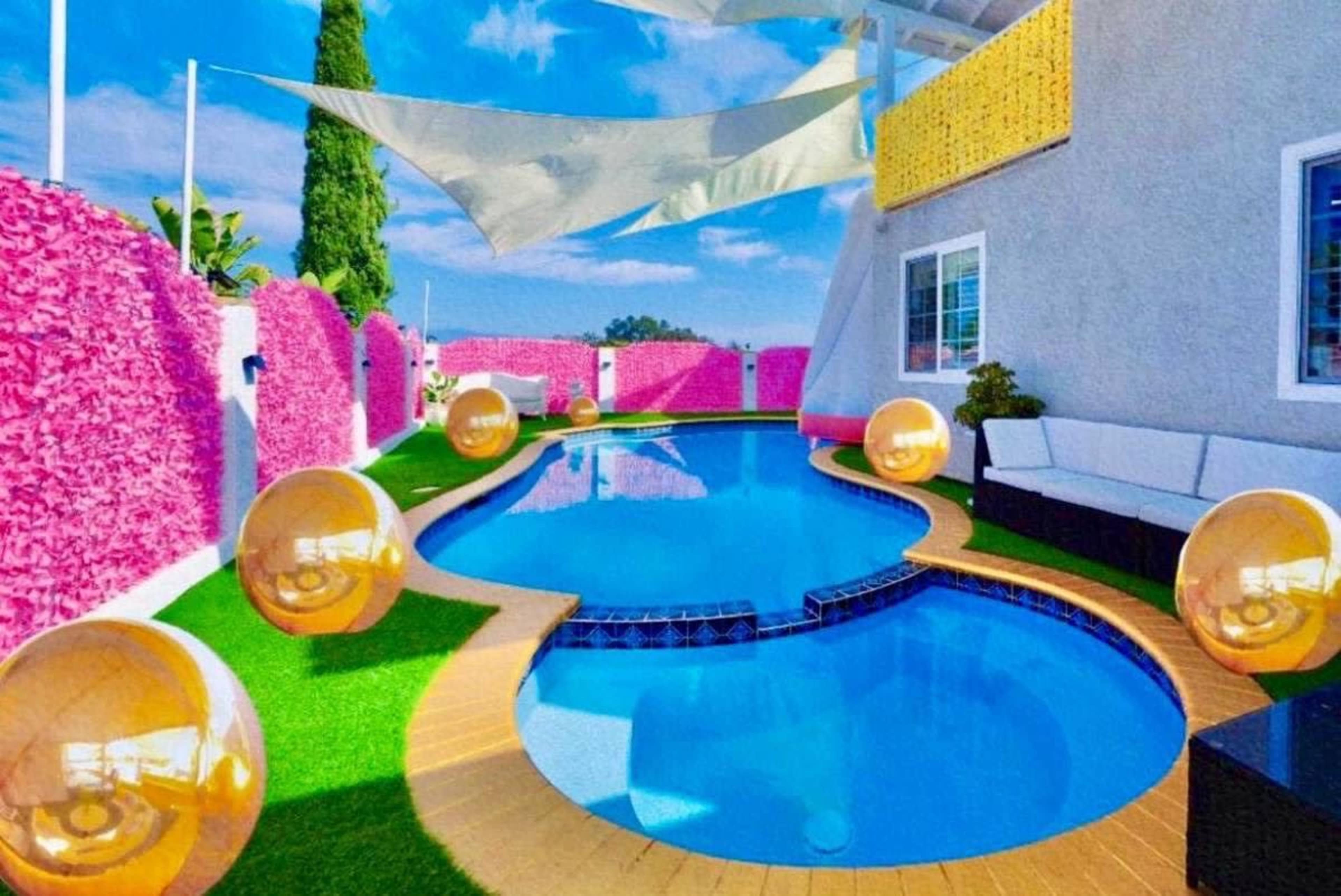 The image shows a vibrant poolside area with a blue swimming pool surrounded by artificial grass and decorative pink walls, complemented by golden spherical ornaments.