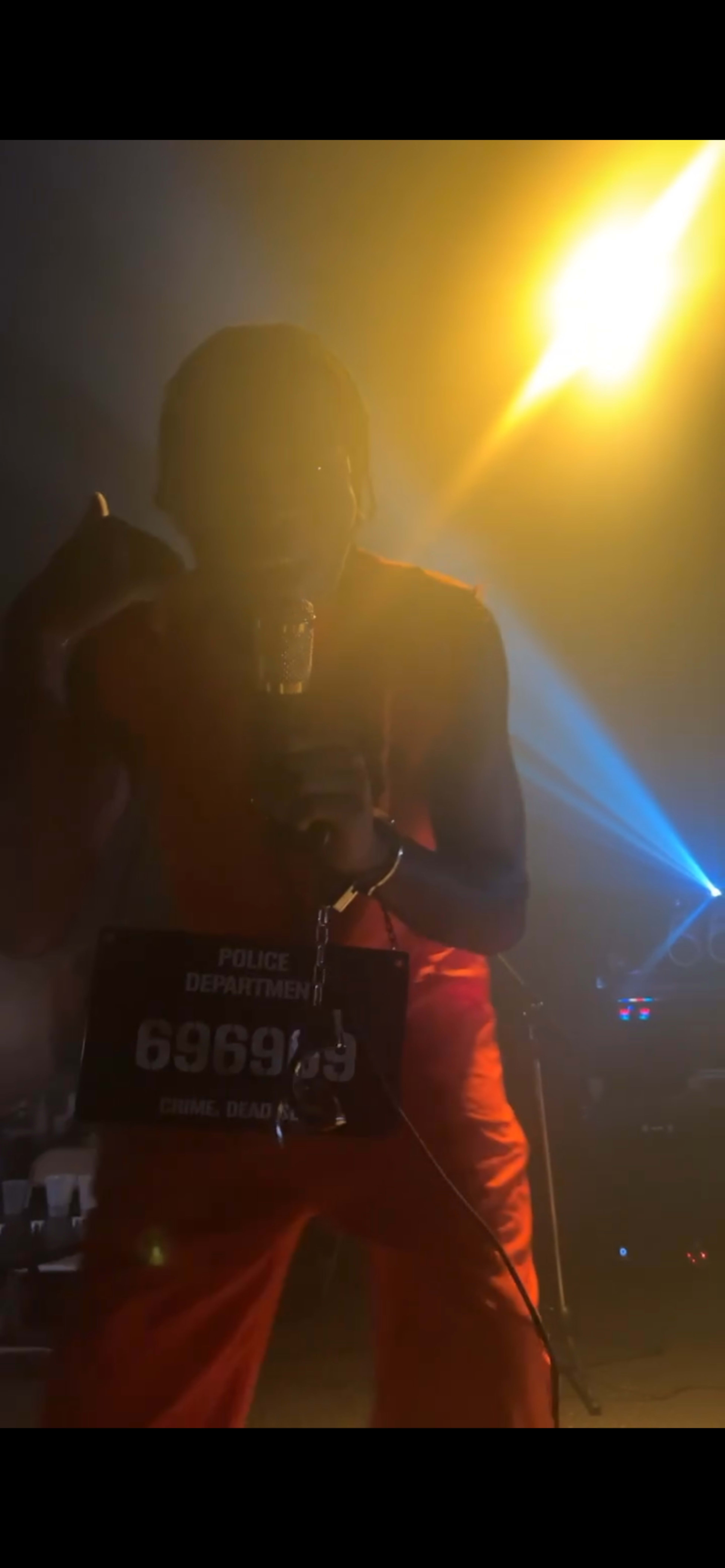 A performer dressed in an orange prison outfit makes a phone gesture while holding a microphone, with theatrical lighting and a police badge number visible.