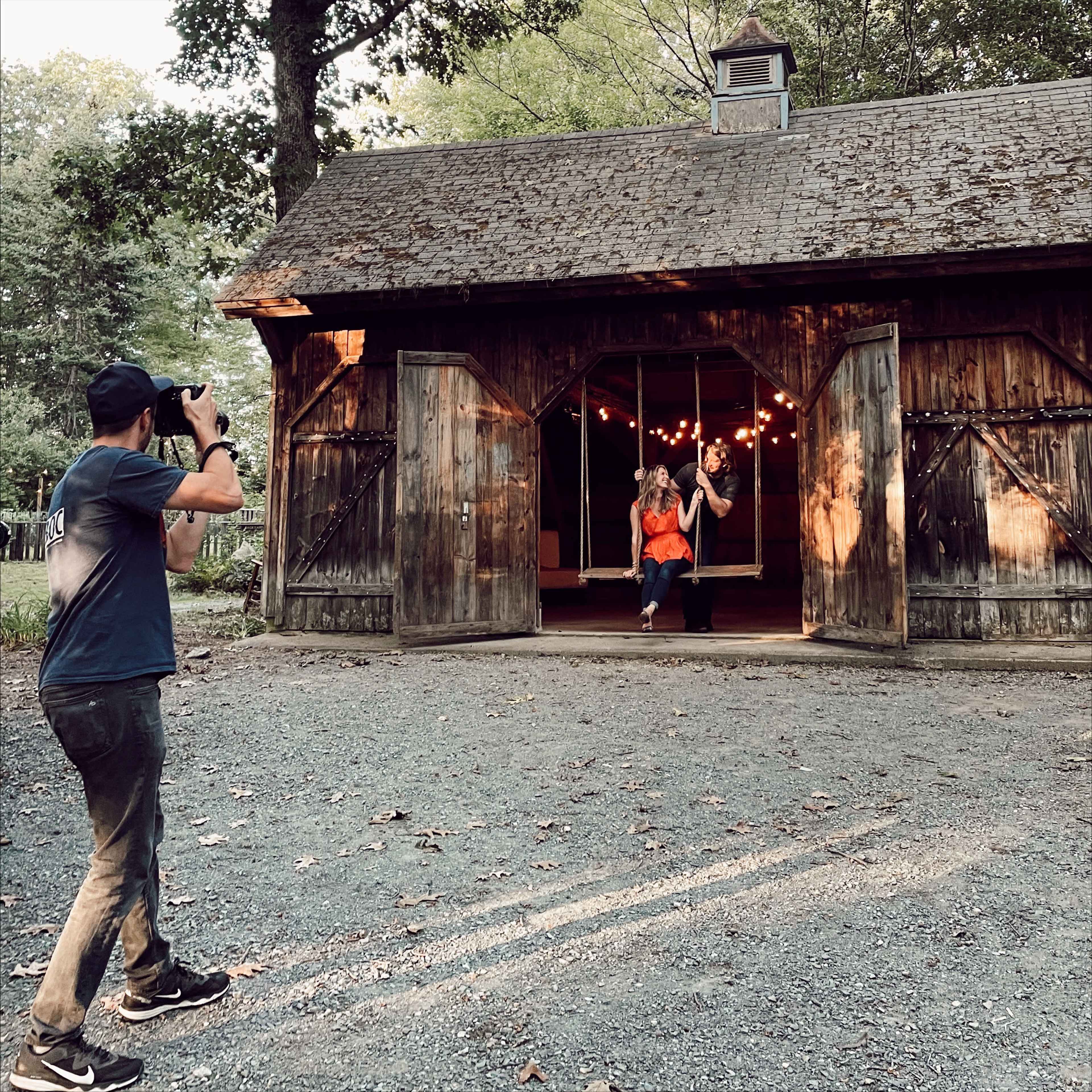 A man takes photographs of two women posing in the doorway of a rustic wooden barn illuminated by string lights.