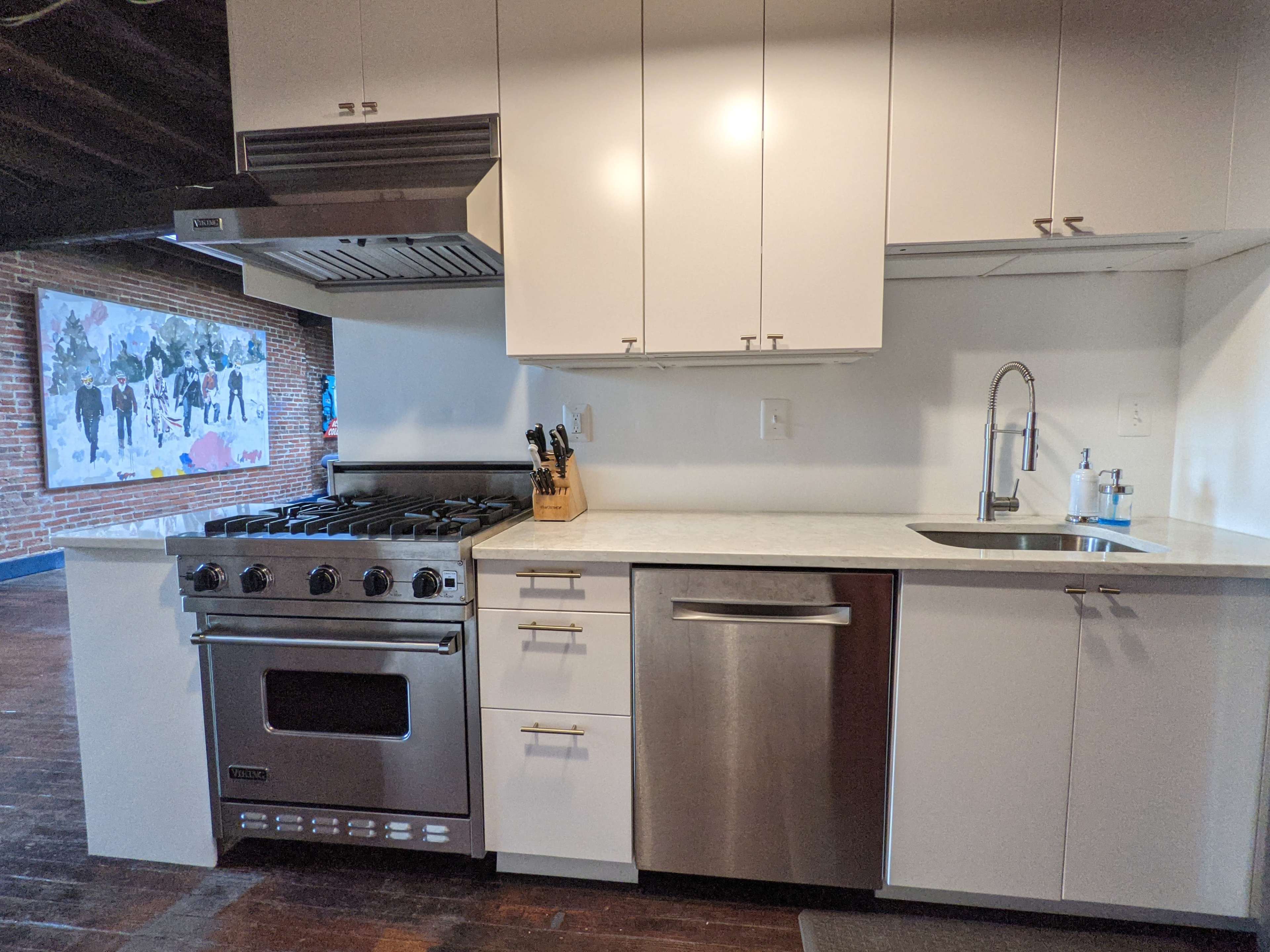 The image shows a modern kitchen with stainless steel appliances, including a gas range and dishwasher, set against a backdrop of exposed brick and a colorful mural.