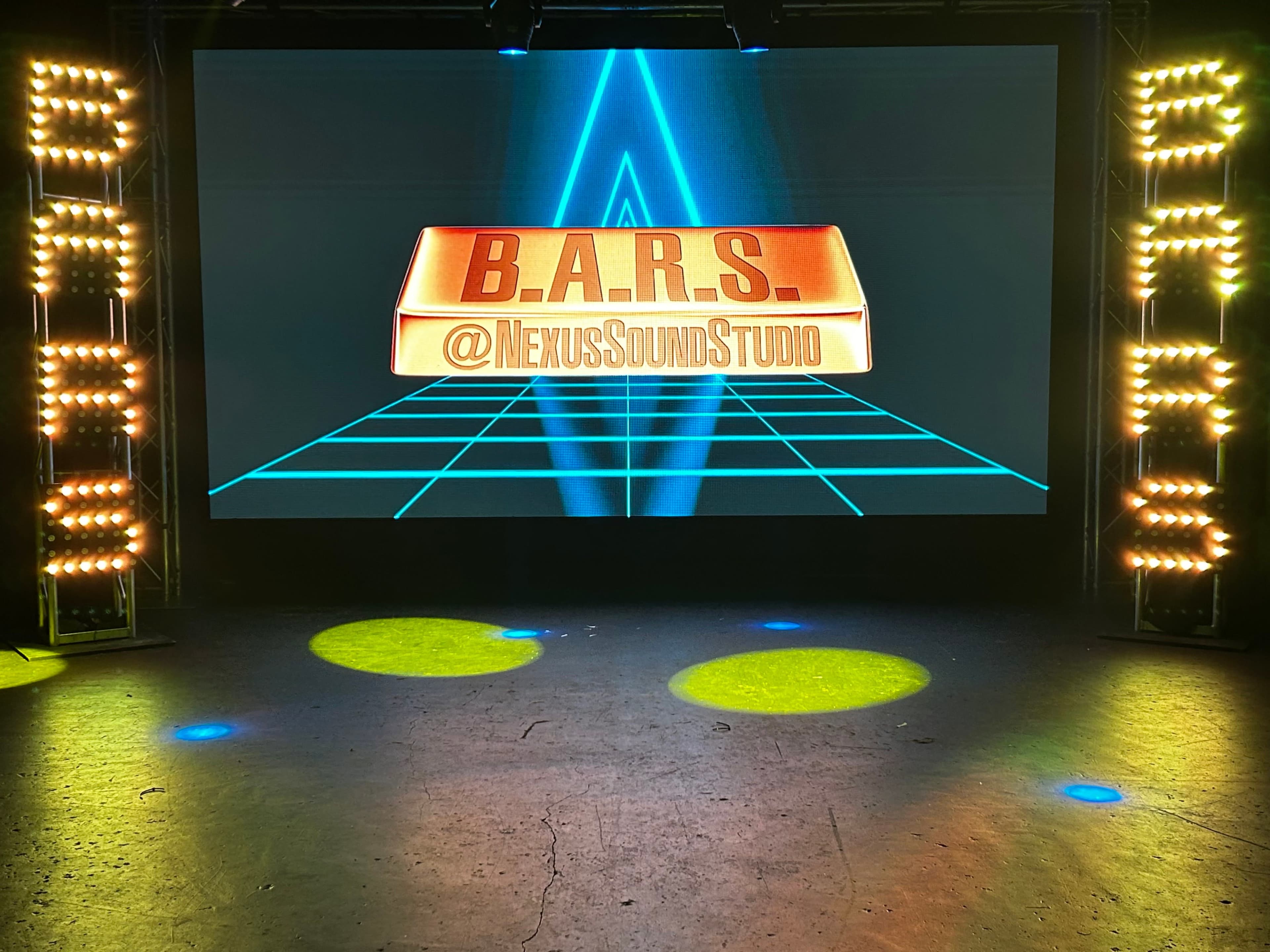 The image shows a stage setup with a large screen displaying "B.A.R.S." and "@NexusSoundStudio," surrounded by lighting fixtures on each side.