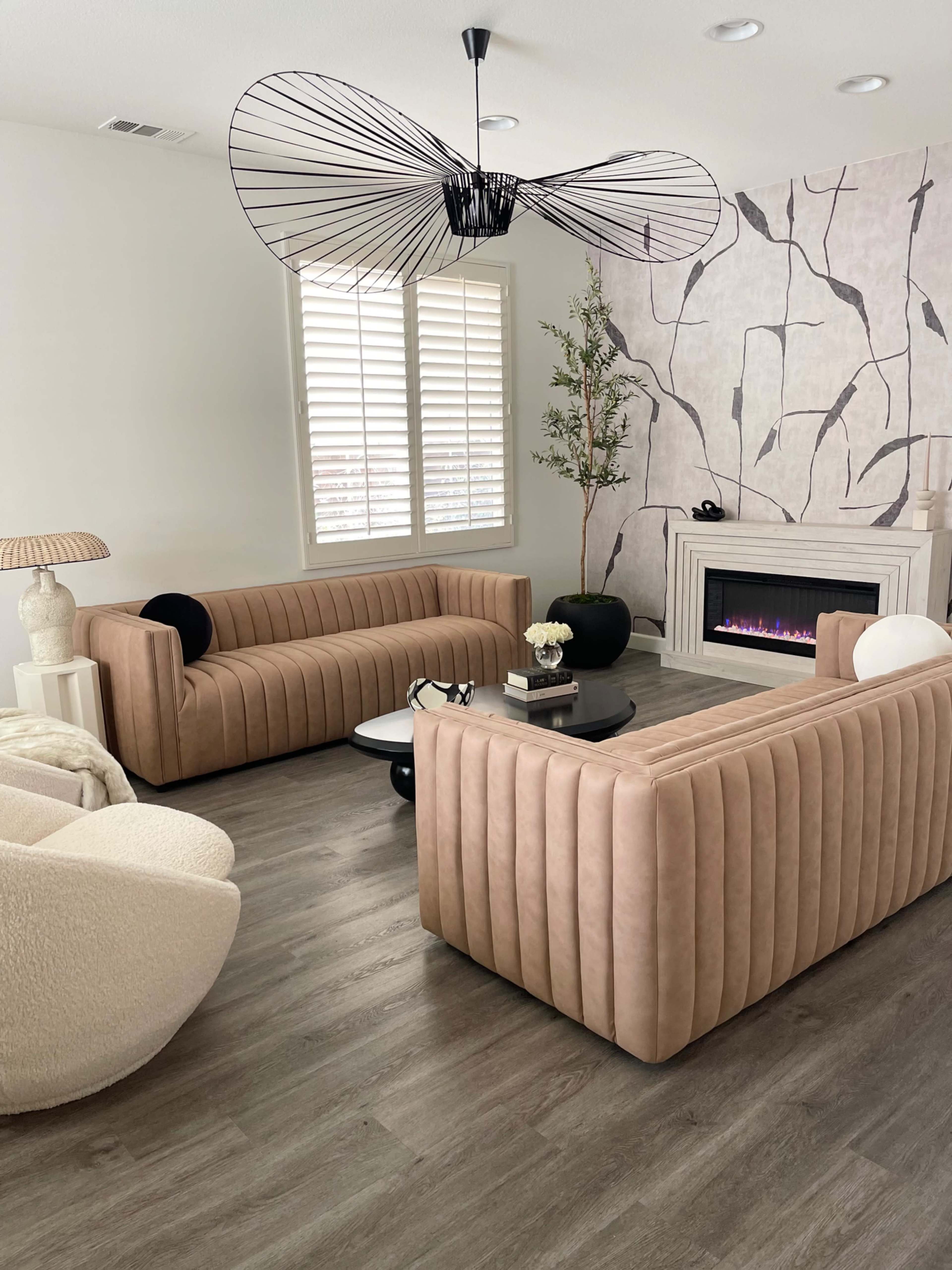 A modern living room features two tufted beige sofas facing each other, accompanied by a round black coffee table and a cozy armchair, with a decorative wall and a gas fireplace in the background.