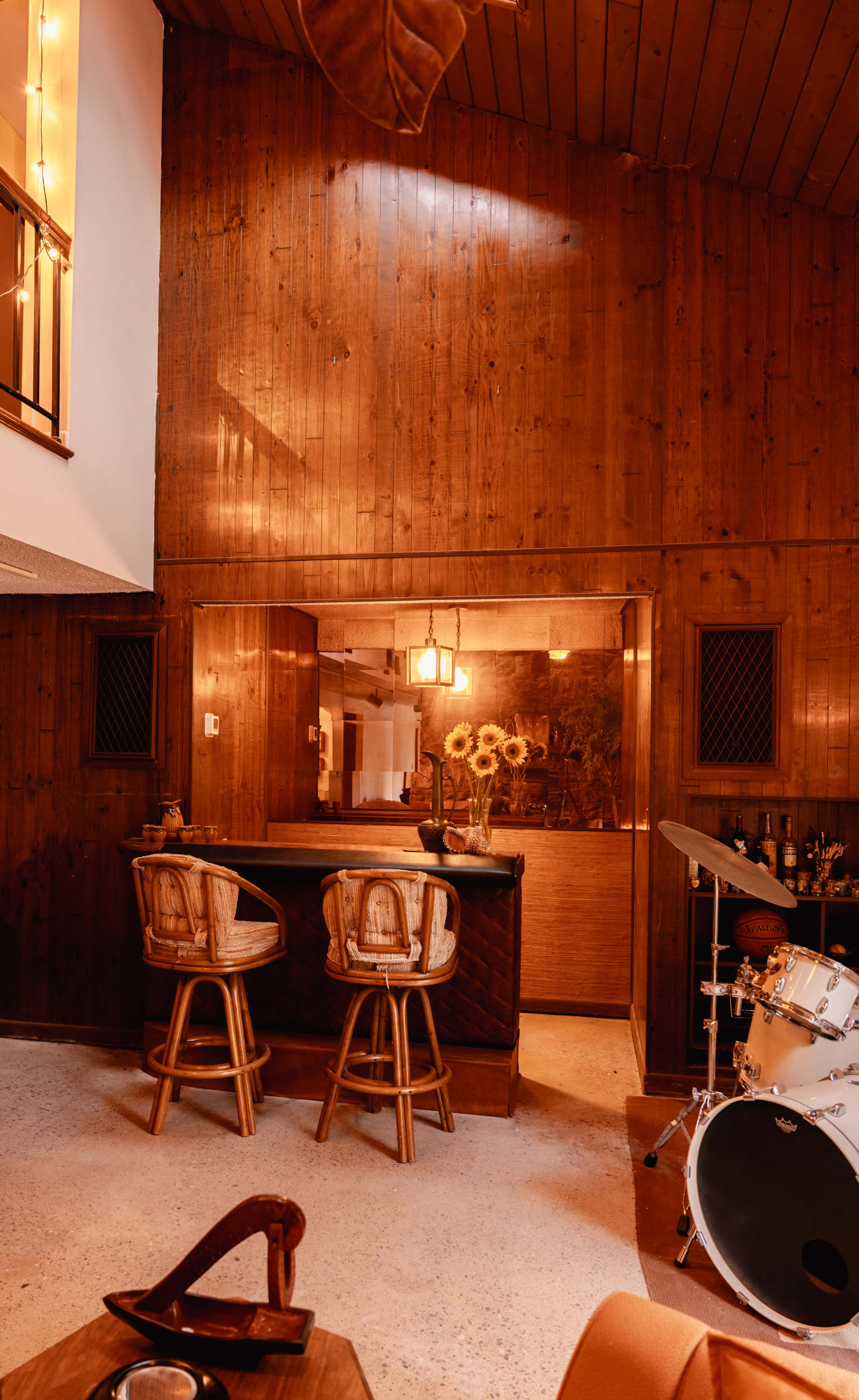 The image shows a cozy interior space featuring a wooden bar with two high chairs, a drum set in the foreground, and soft lighting illuminating the room.