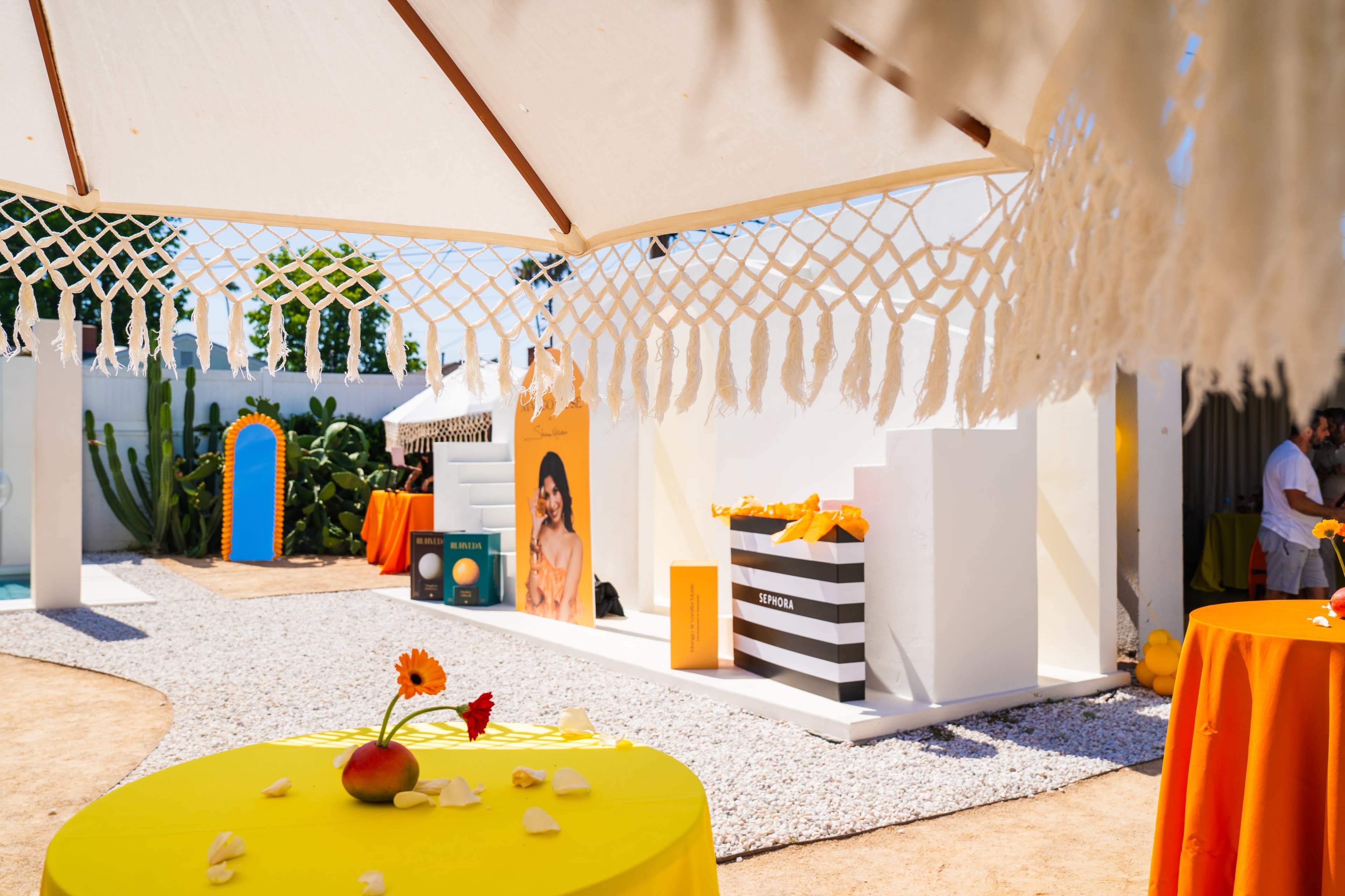 The image shows a vibrant outdoor event space with yellow tables, decorative elements, and a backdrop featuring promotional displays.