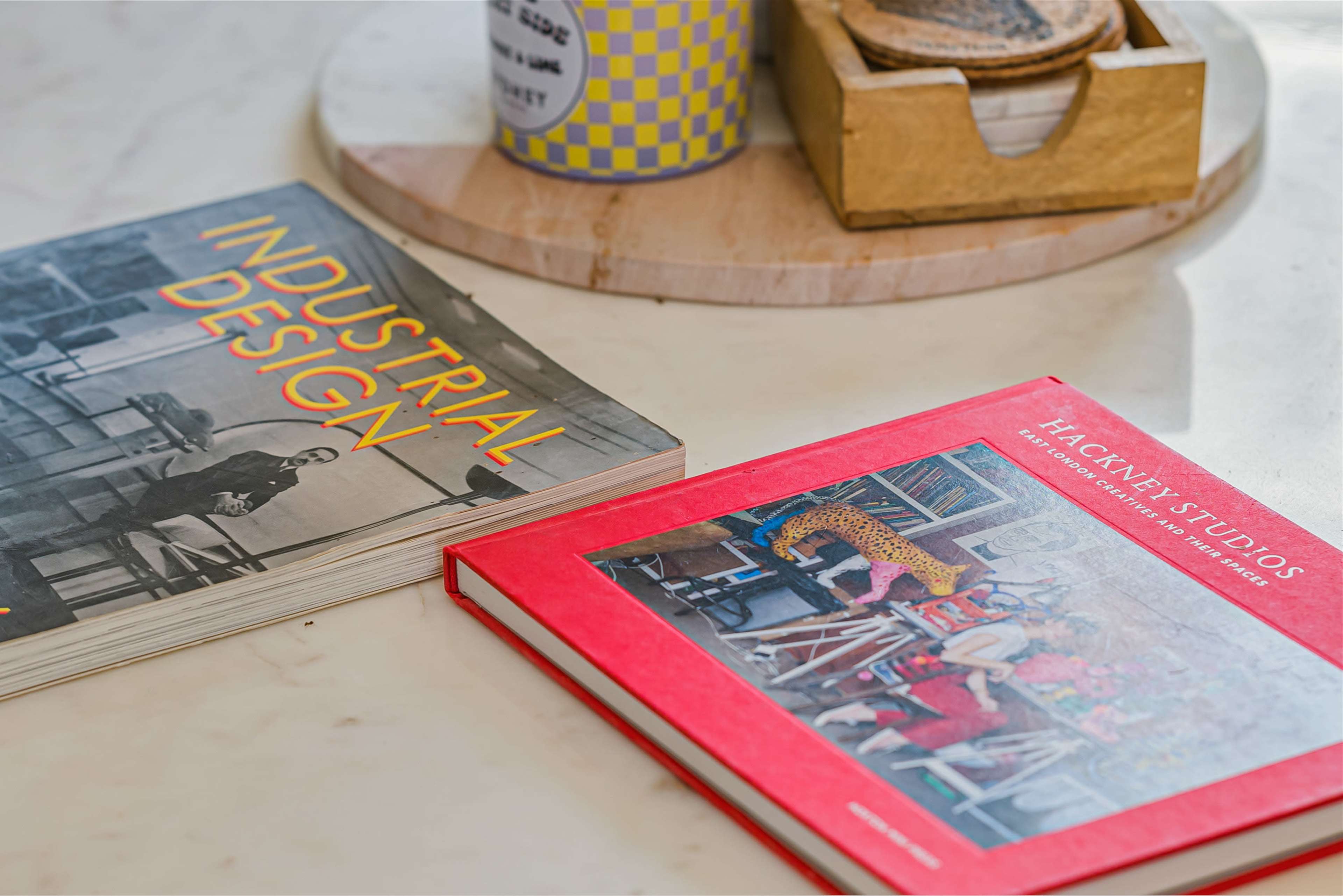 The image features two books, one titled "Industrial Design" and the other "Hackney Studios," placed on a round wooden tray next to a mug and a small container.
