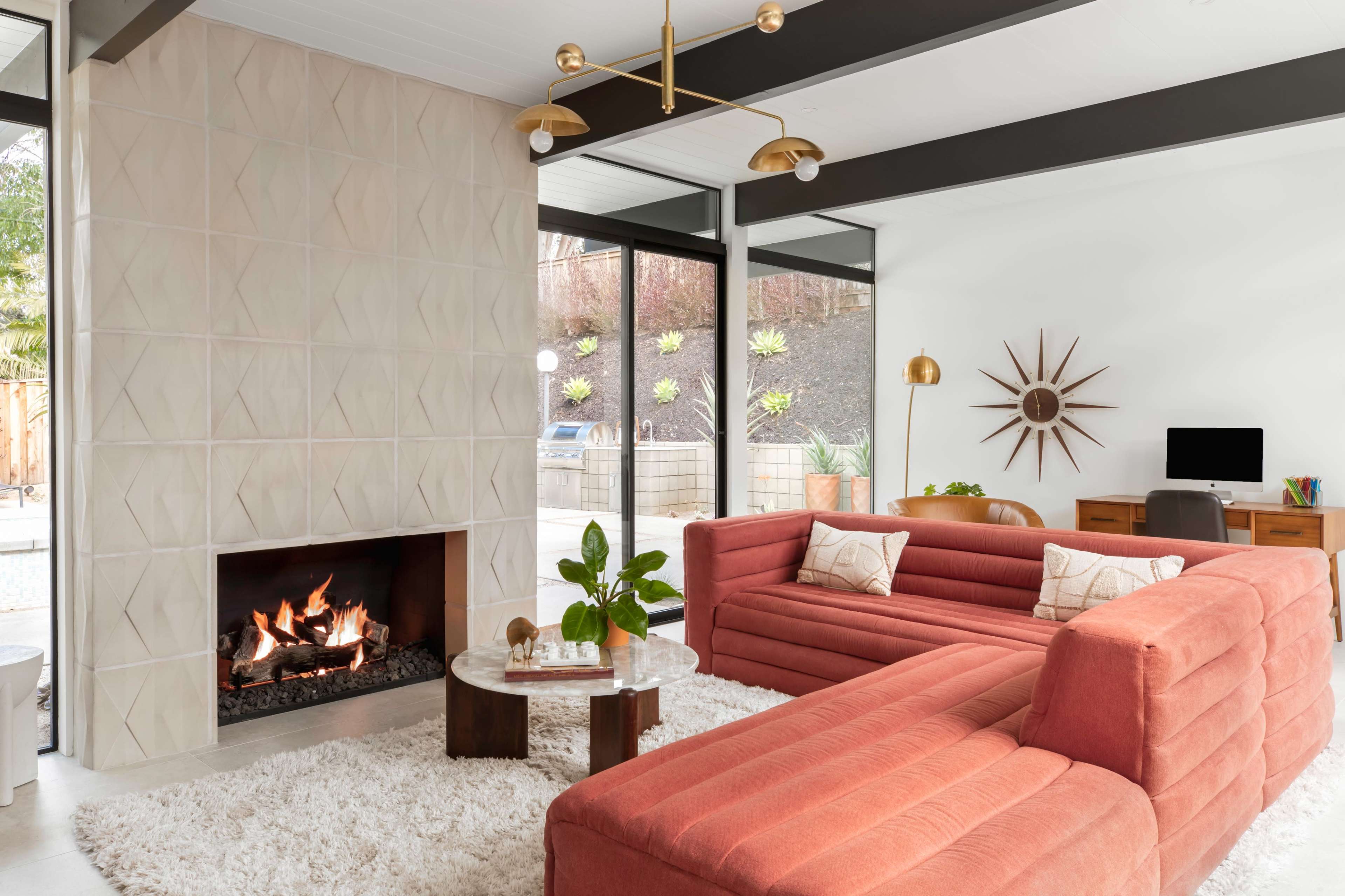 A mid-century modern living room features a coral sectional sofa, a circular coffee table, a fireplace, and a workspace with a computer, all surrounded by large windows.