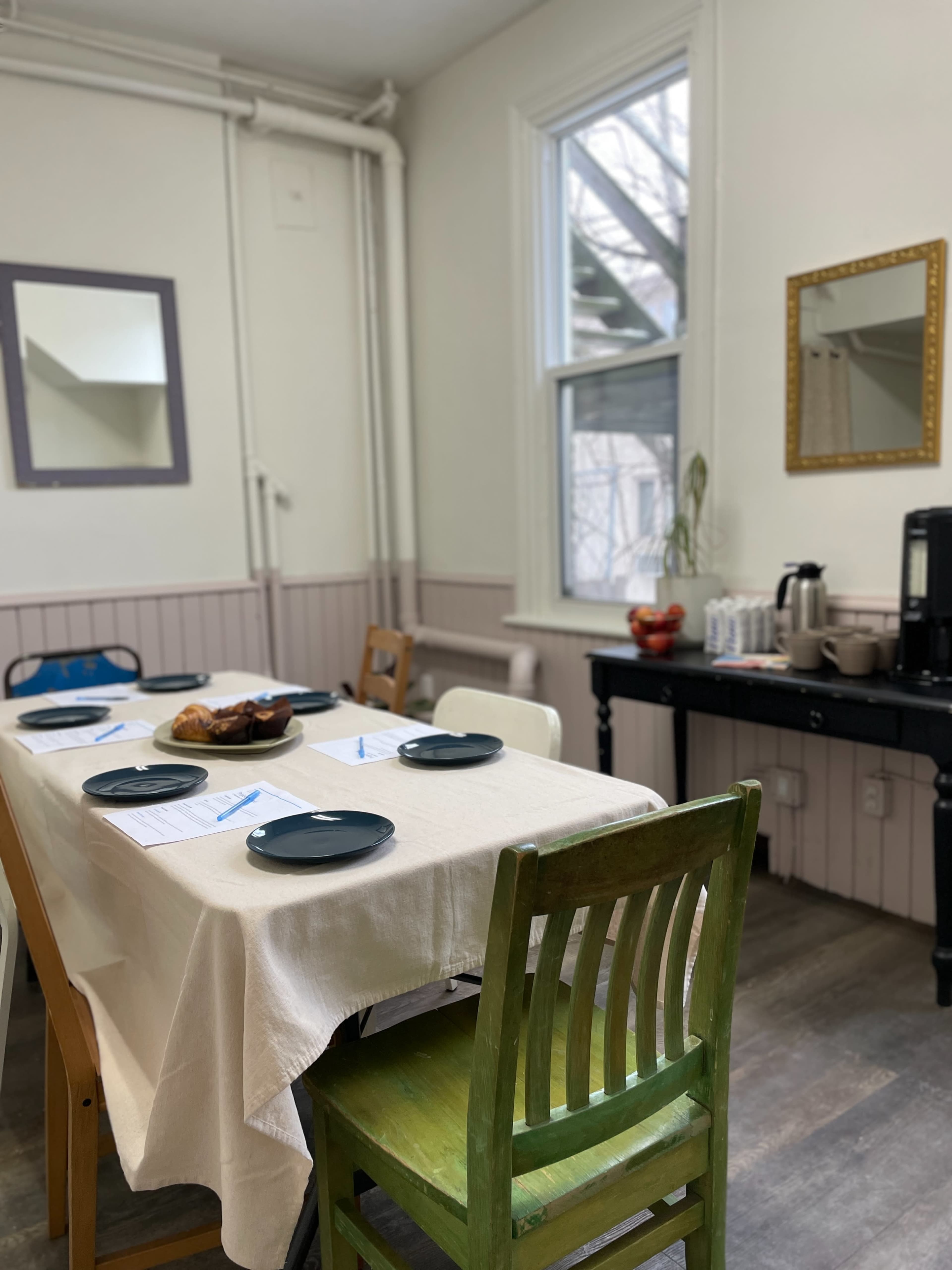 A dining area features a table set with plates and a vase, surrounded by several chairs and a side table with a coffee maker and snacks.
