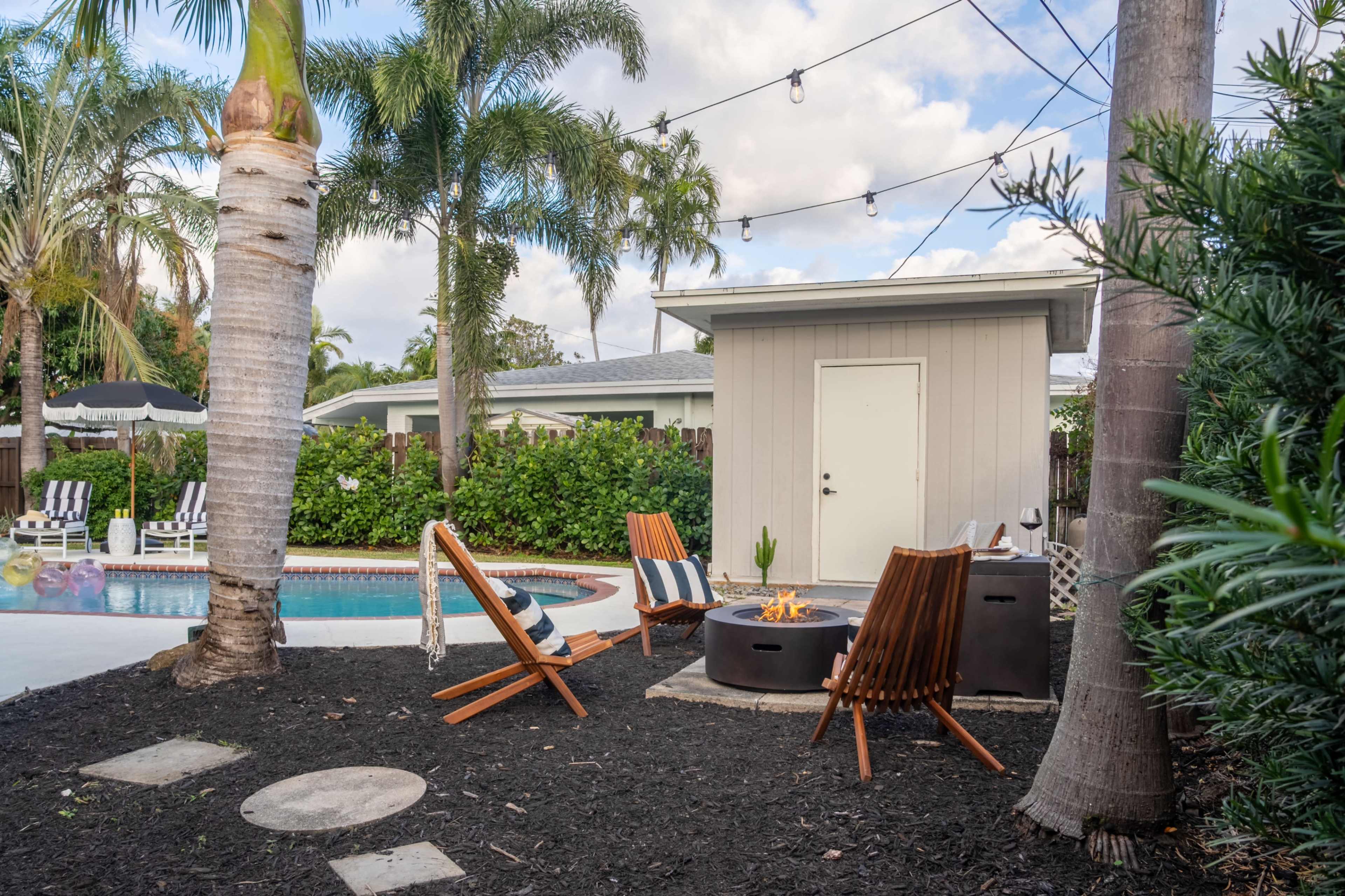A small backyard features a pool surrounded by lounge chairs, a fire pit with seating, and a shed in the background, all bordered by palm trees.