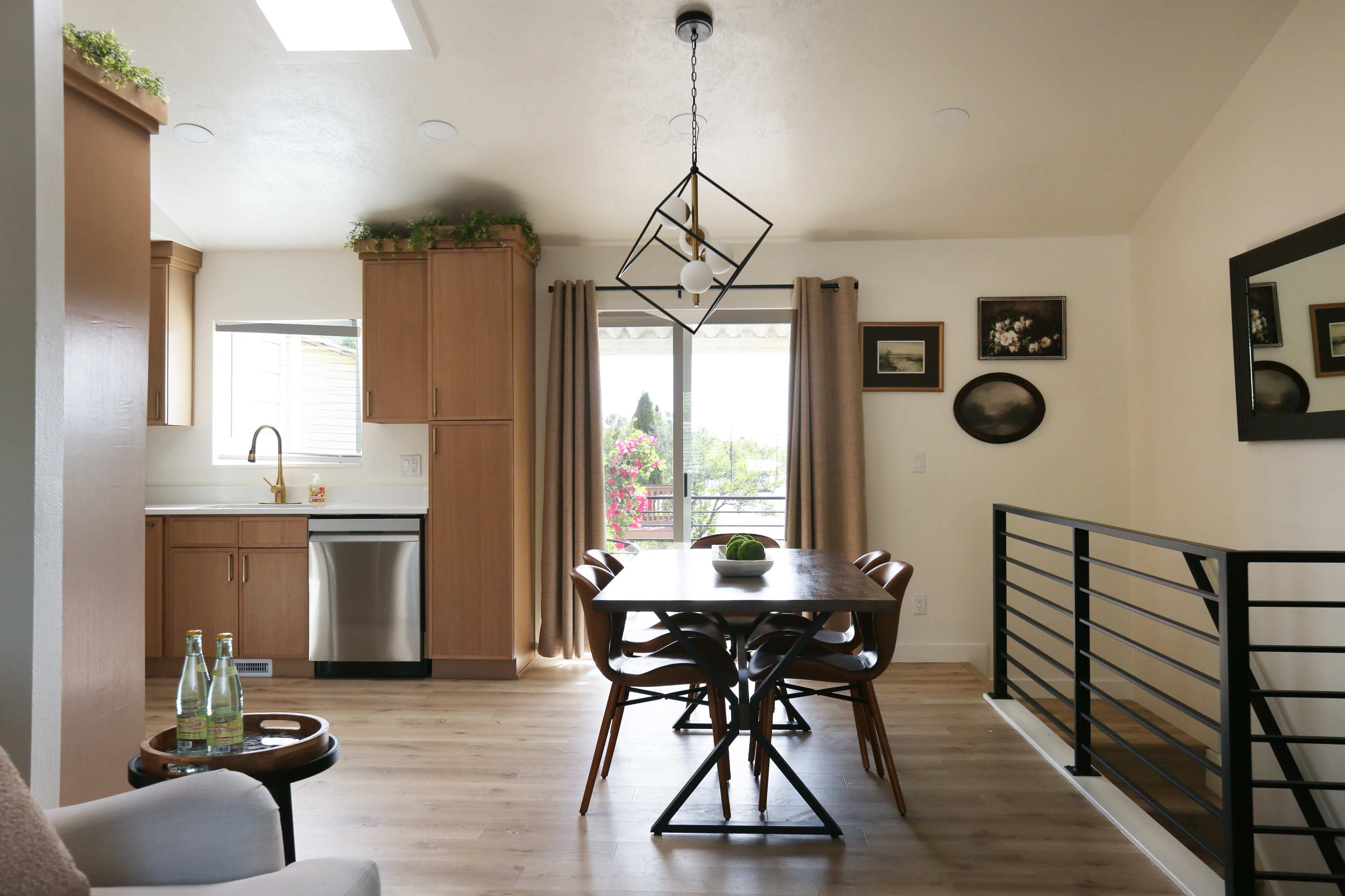 The image depicts a modern dining area with a wooden table surrounded by chairs, a kitchen in the background, and large windows letting in natural light.
