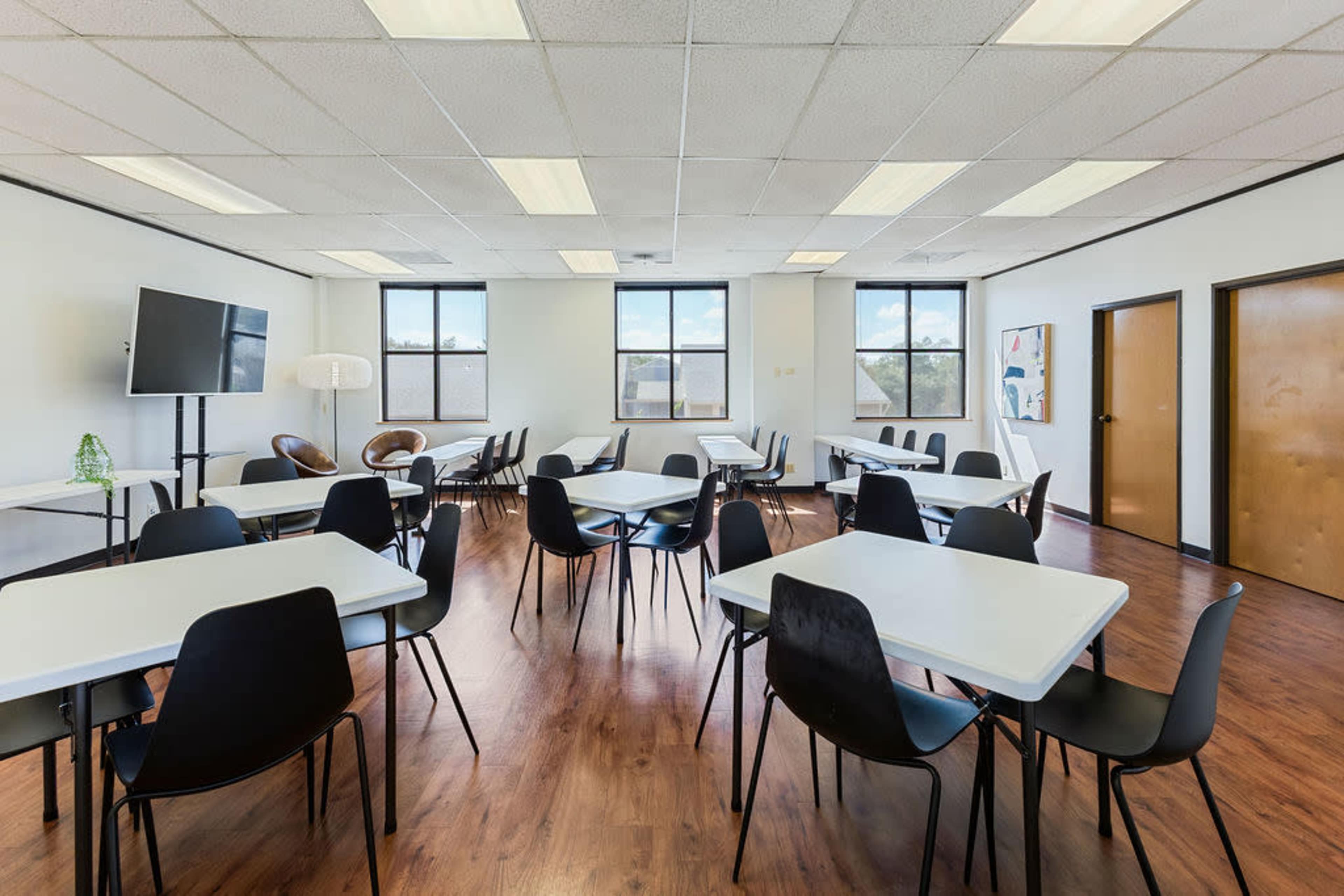 The image shows a spacious classroom or meeting room filled with several white tables and black chairs, with large windows allowing natural light to enter.