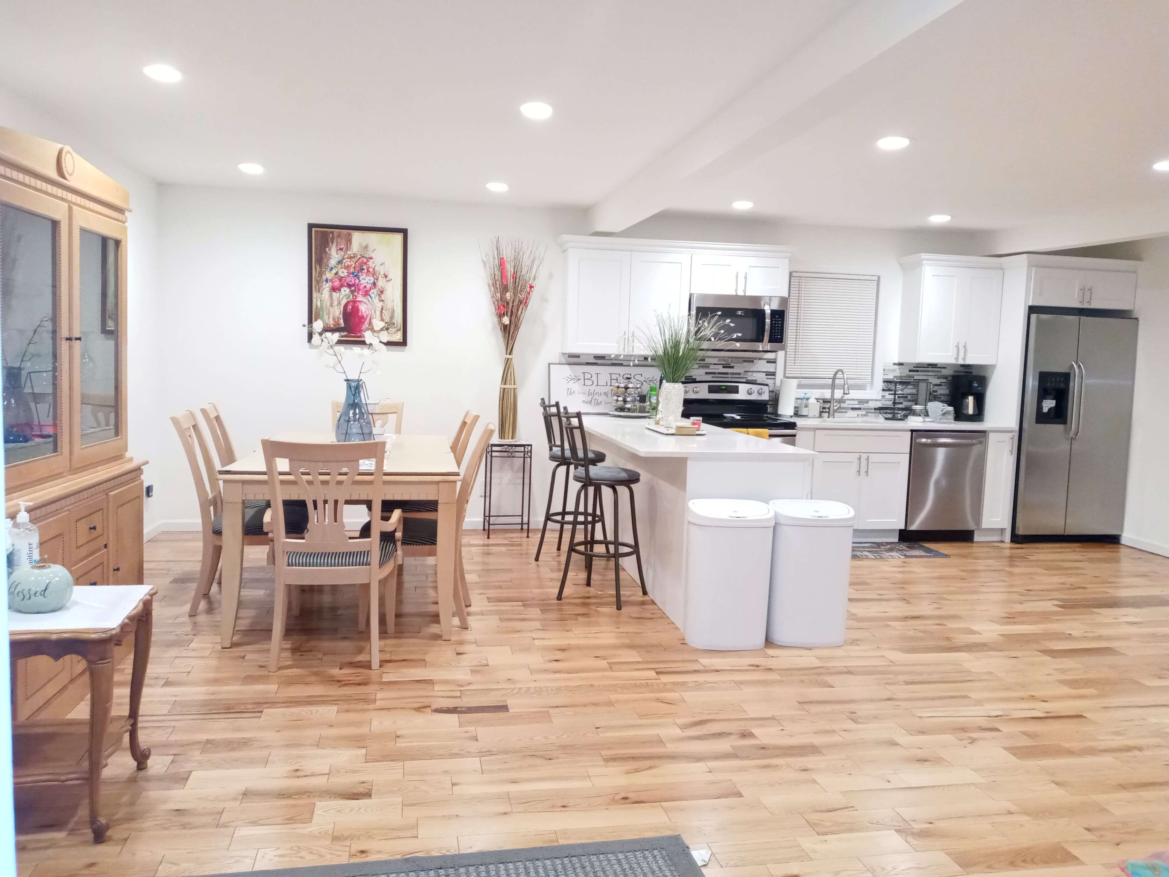 A modern kitchen and dining area with wooden flooring, white cabinetry, a dining table set for six, and a stainless steel refrigerator.