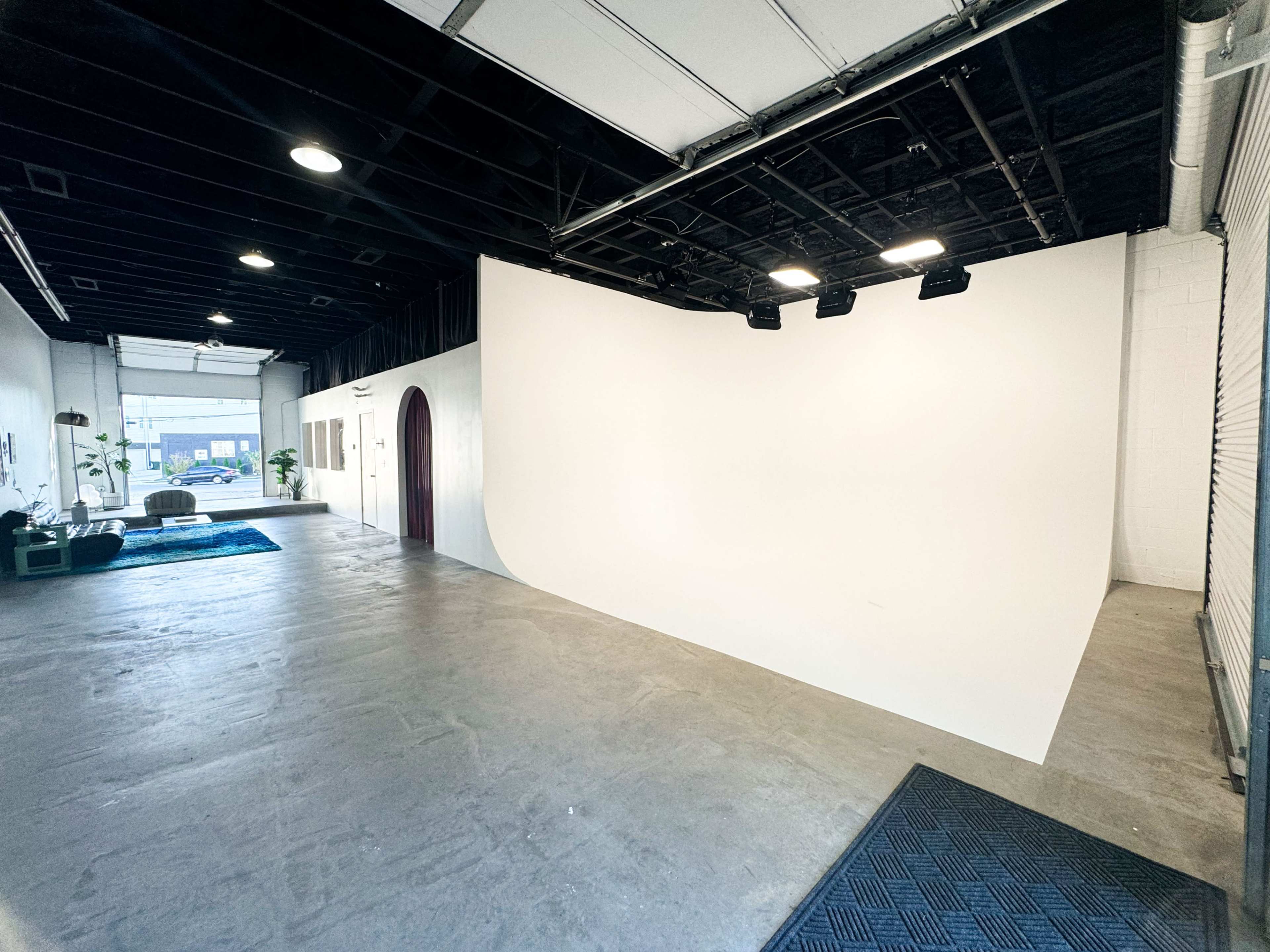 The image shows a spacious studio with a smooth white curved wall, concrete flooring, and a large entrance area illuminated by overhead lights.