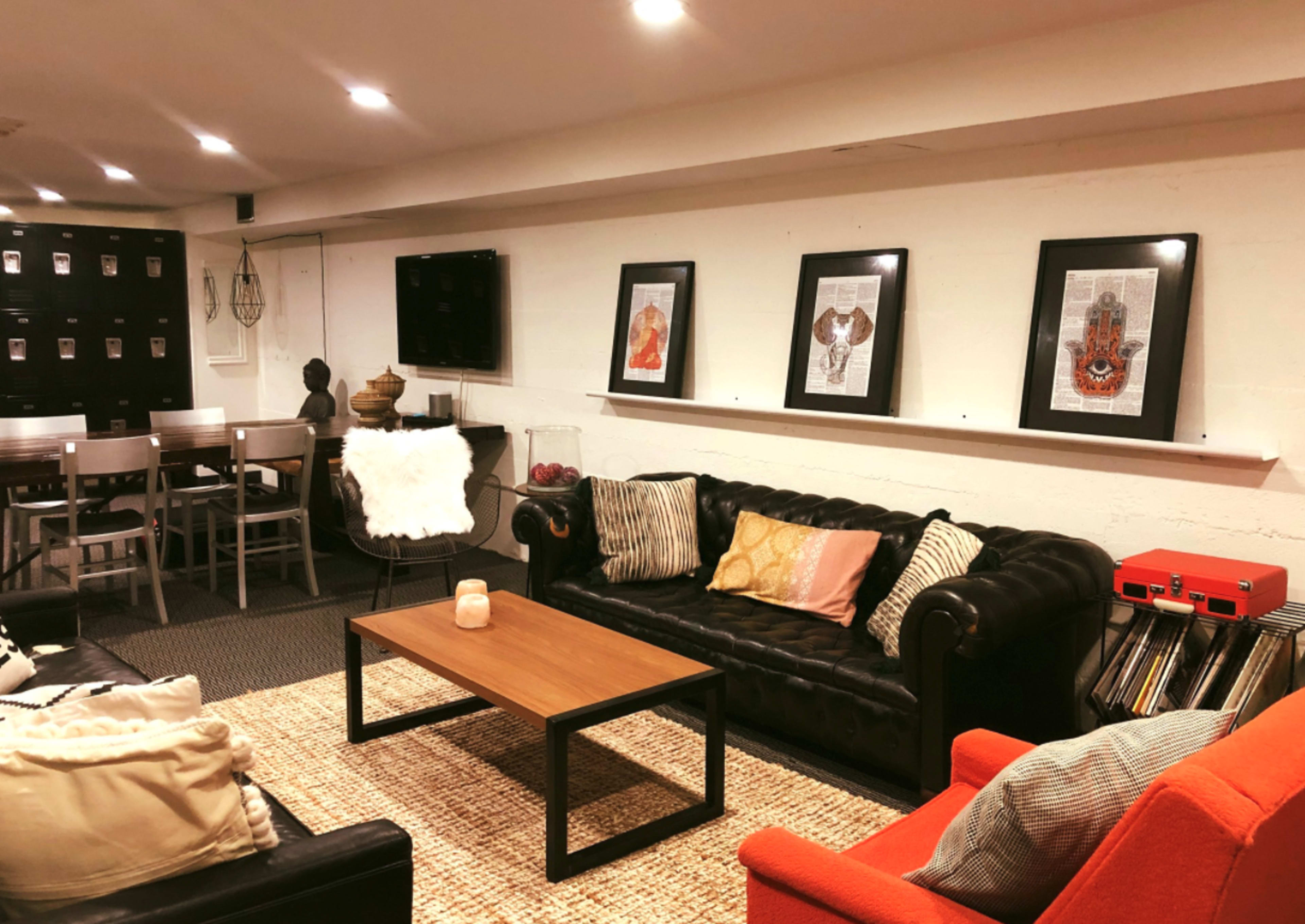 A cozy basement lounge area with a black leather sofa, an orange chair, a wooden coffee table, and framed artwork on the wall.