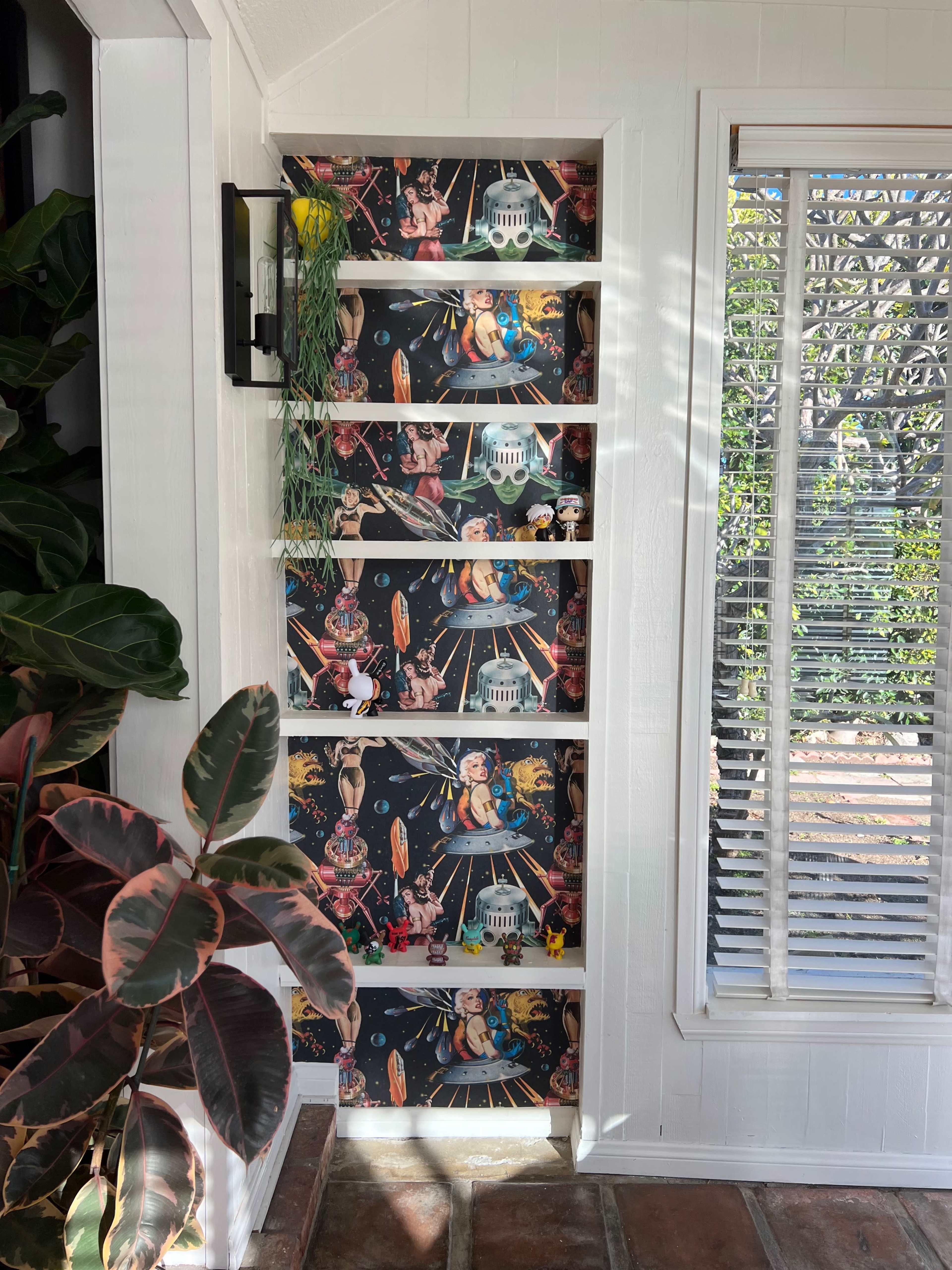 A narrow shelving unit against a wall with colorful, patterned wallpaper featuring various whimsical illustrations.