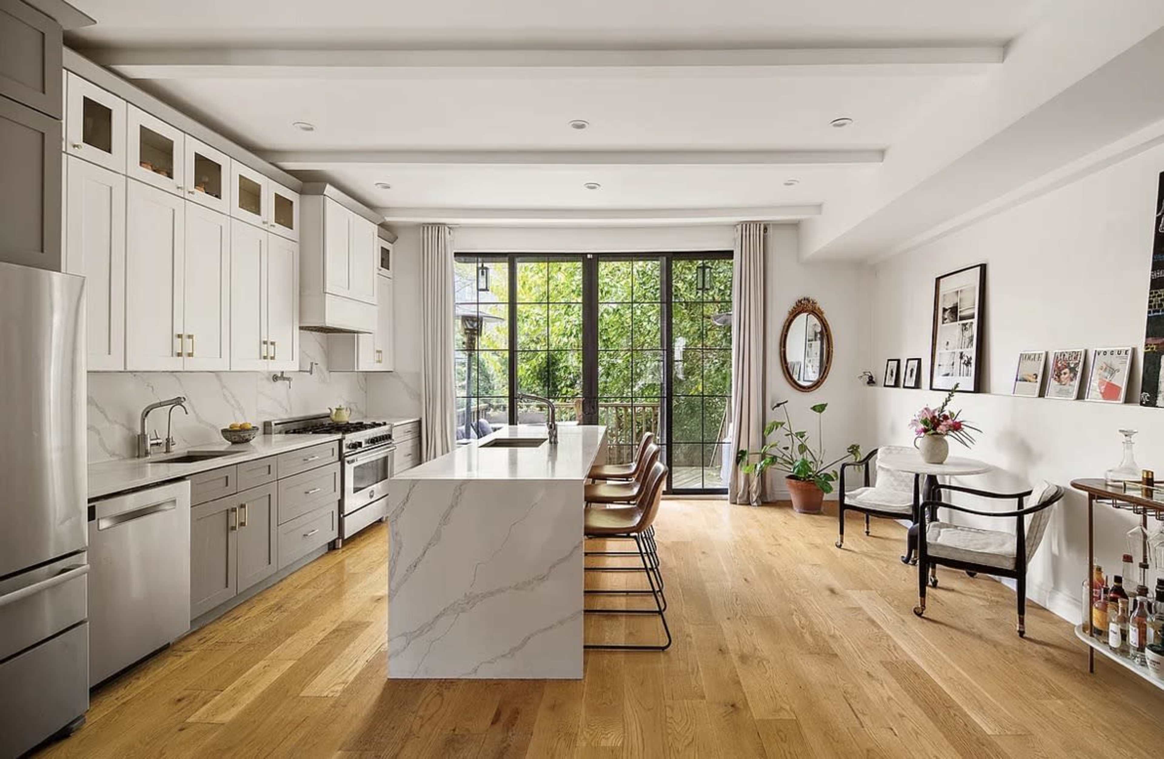 The image shows a modern kitchen with white cabinetry, a large island with a marble top, and large windows showcasing greenery outside.