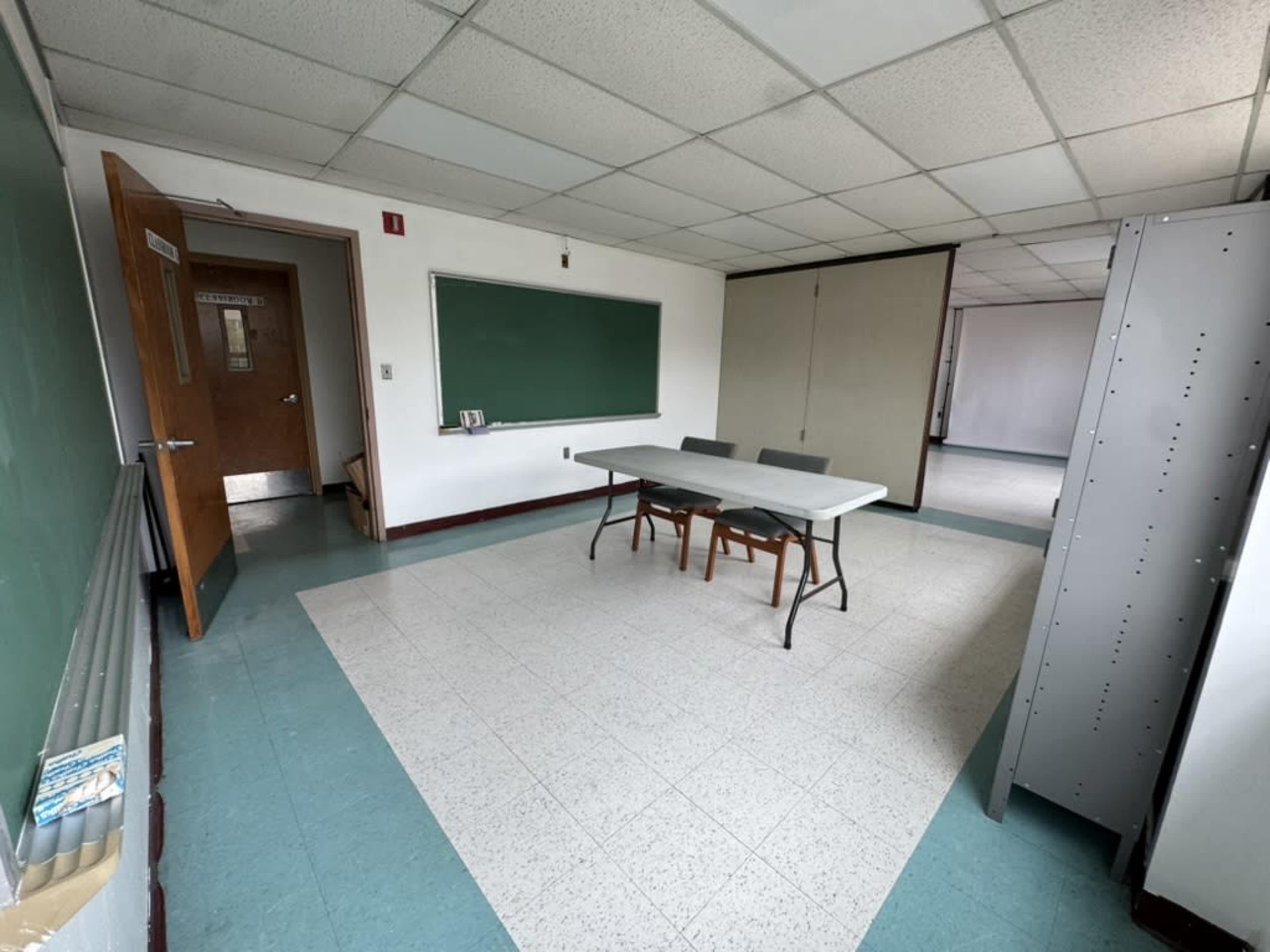 The image shows a sparsely furnished classroom with a green chalkboard, a table with chairs, and a storage unit against the wall.