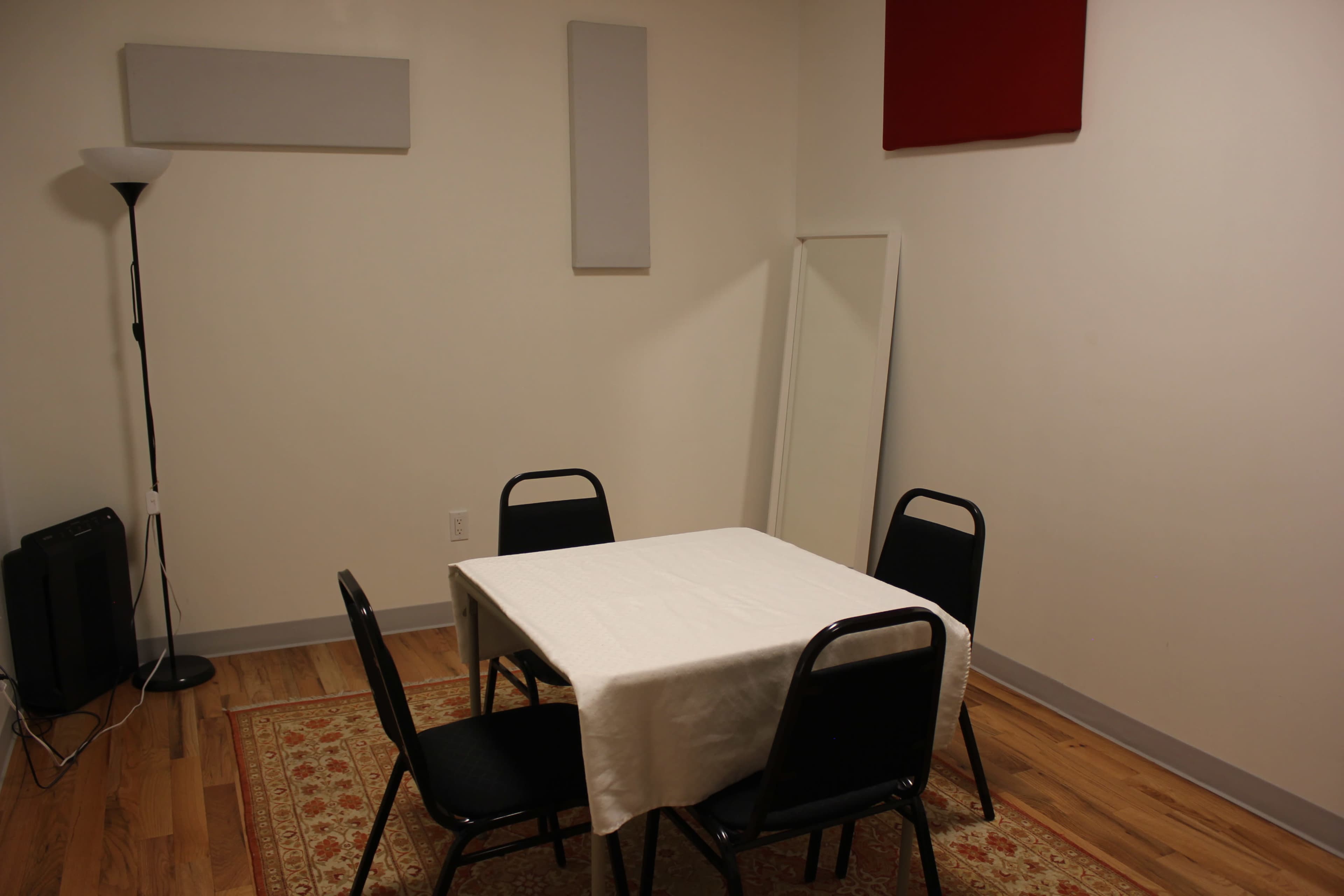 A small room with a table covered by a white cloth and four black chairs arranged around it, along with a floor lamp, a mirror, and soundproofing panels on the walls.