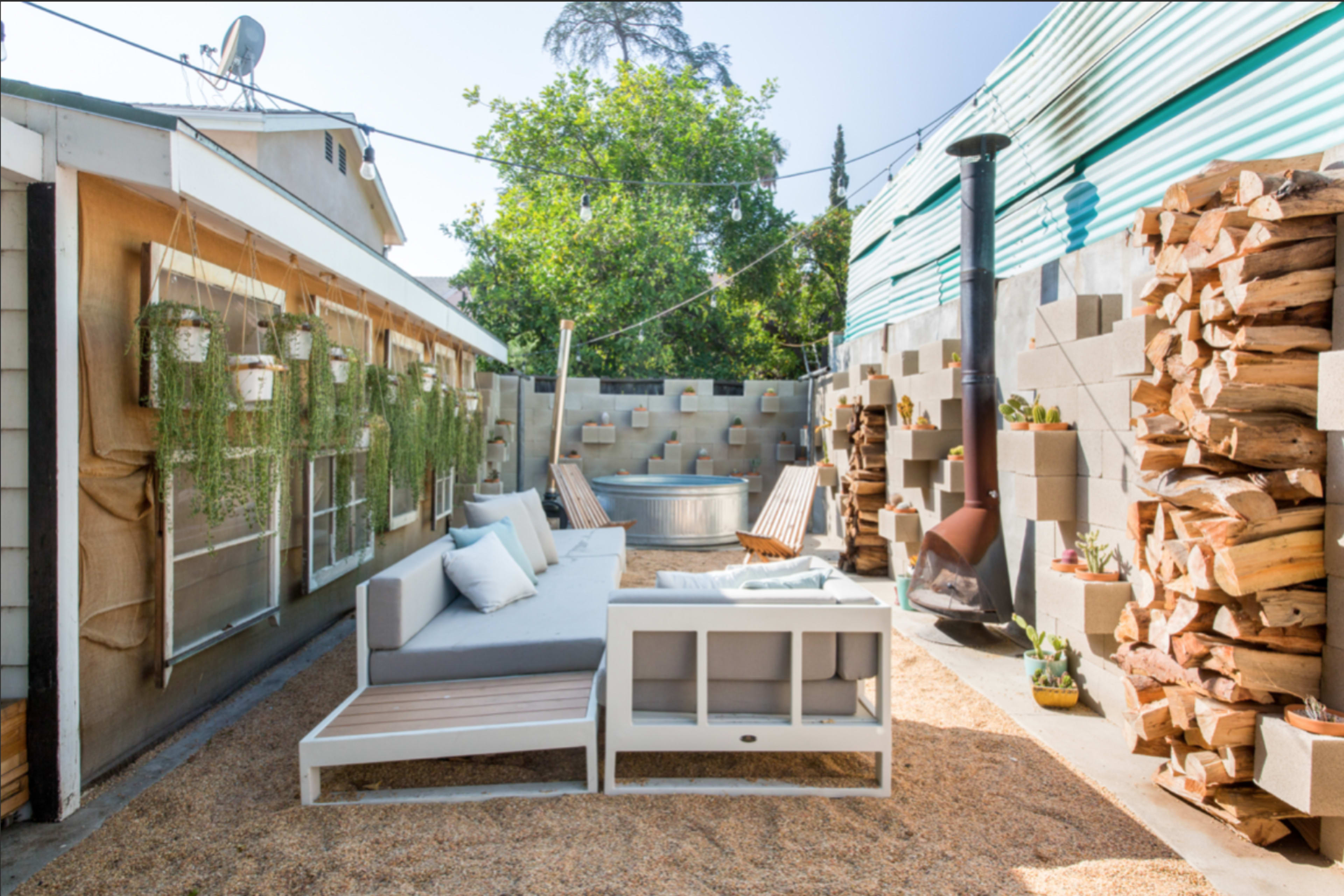 A backyard space featuring a gray sectional sofa, wooden chairs, a small fire pit, and stacked firewood against a textured wall.