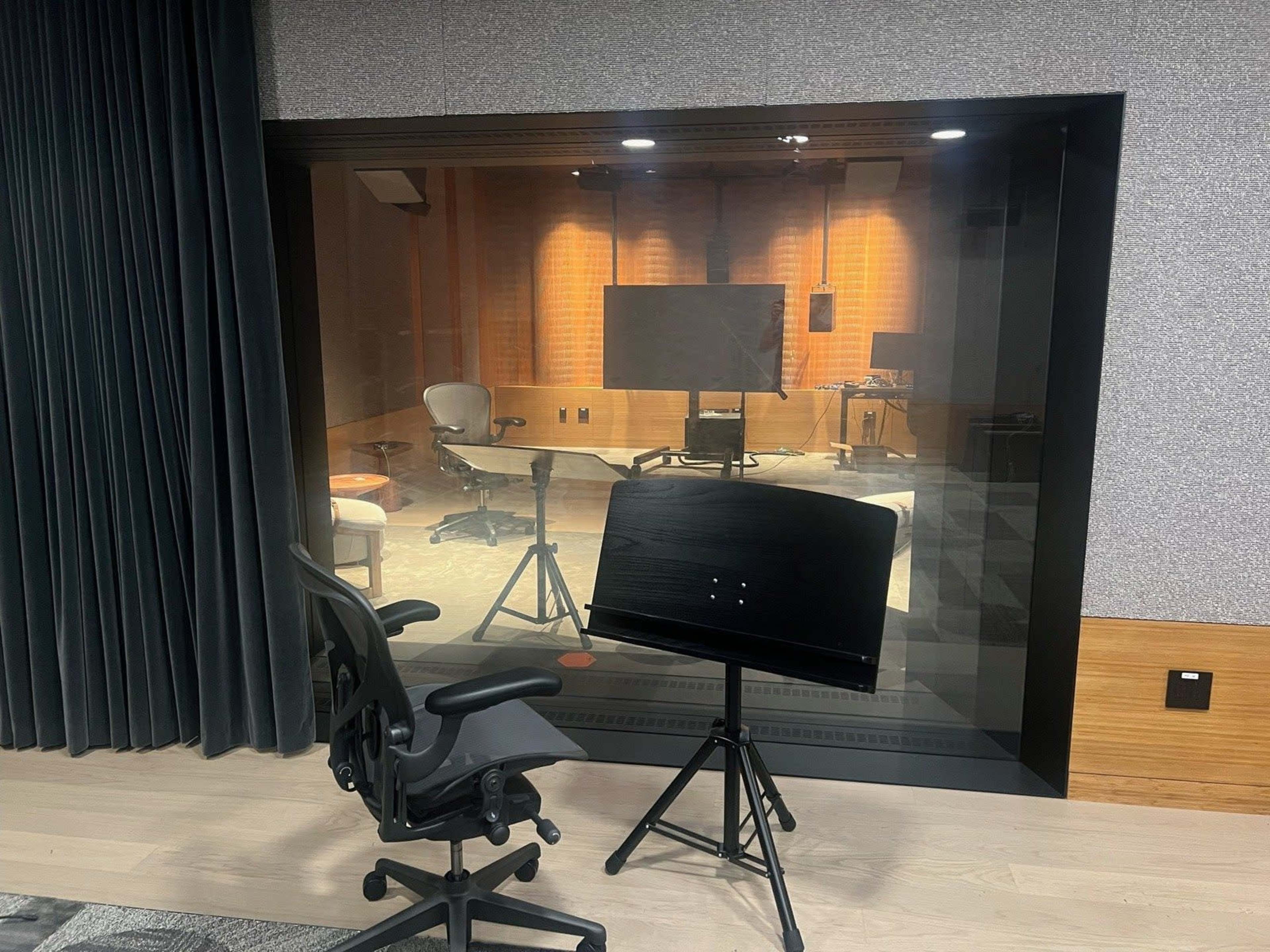 The image shows a modern recording studio with glass walls, featuring a chair, a music stand, and equipment inside the soundproofed area.