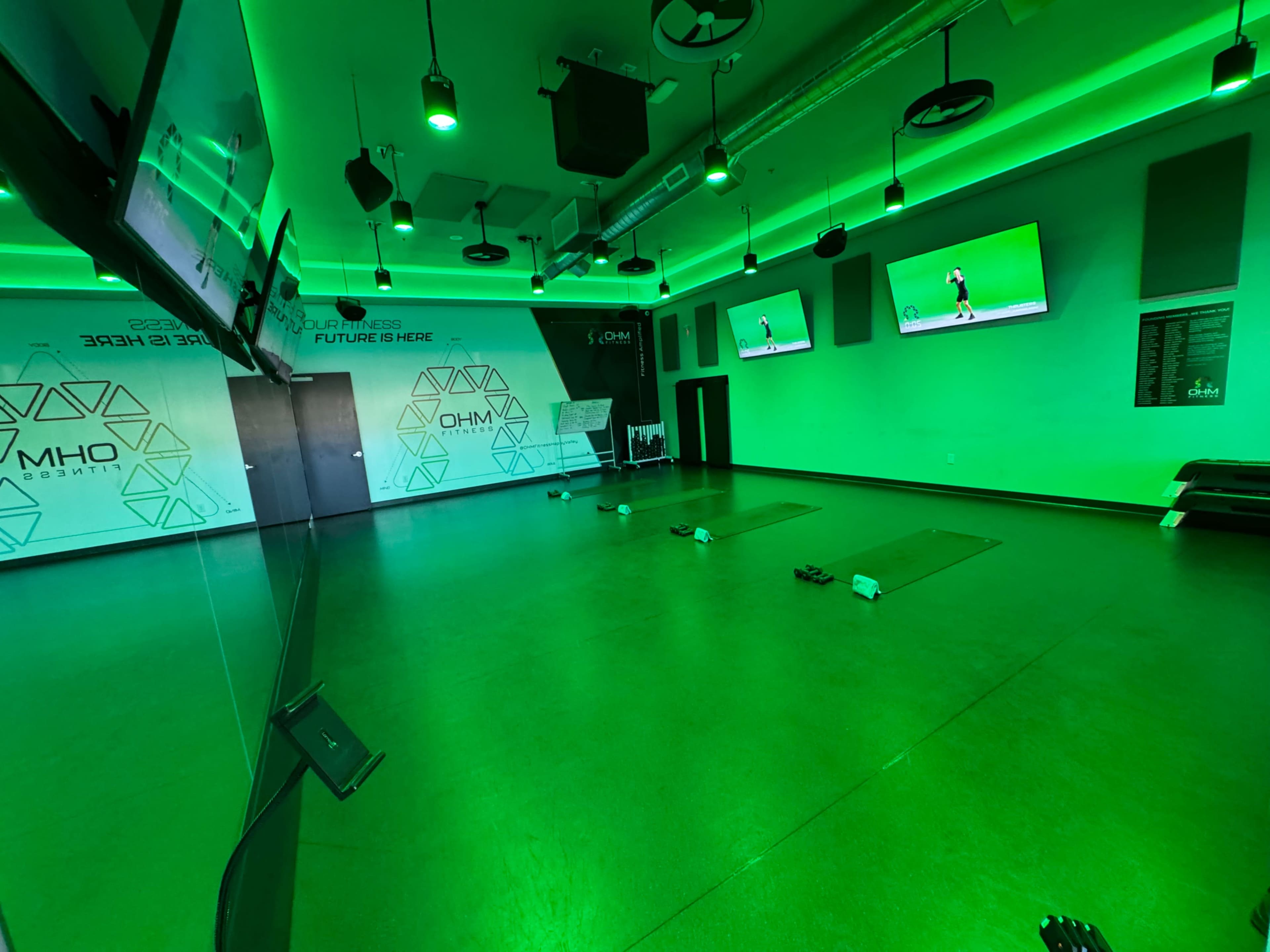 The image shows a fitness studio with green lighting, featuring exercise mats, weights, and multiple screens displaying workout sessions.