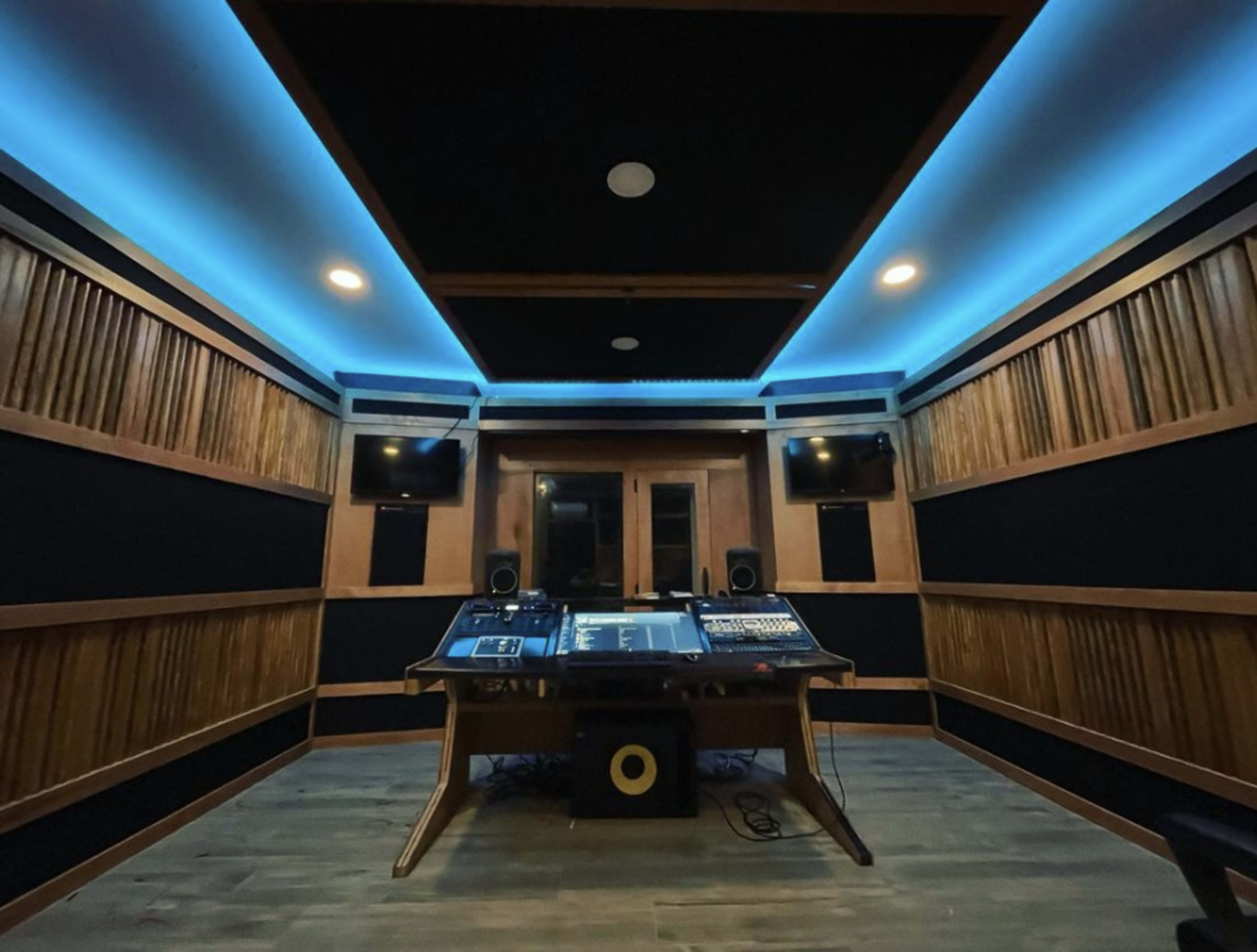 A modern recording studio with wooden walls, a sound mixing desk in the center, and blue LED lights illuminating the ceiling.