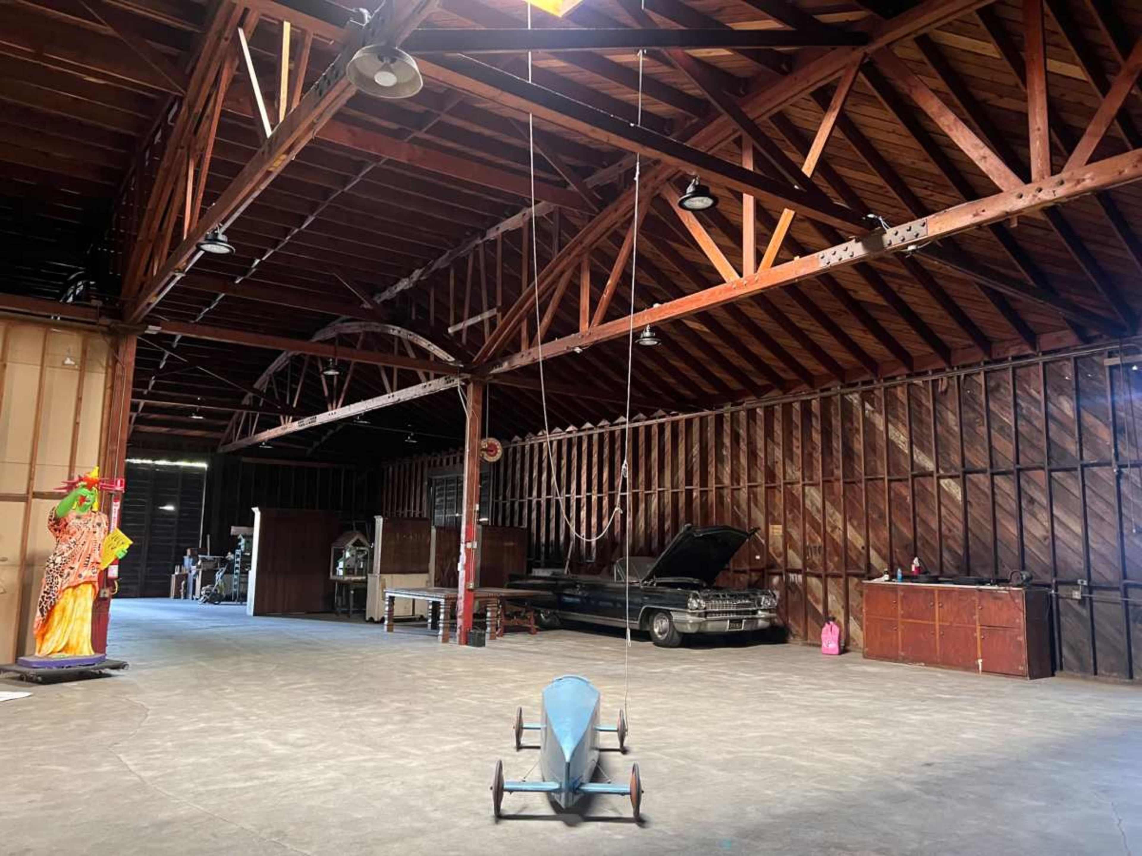 The image shows the interior of a large, empty barn with exposed wooden beams, an old car parked in the corner, and a colorful decorative figure in the foreground.