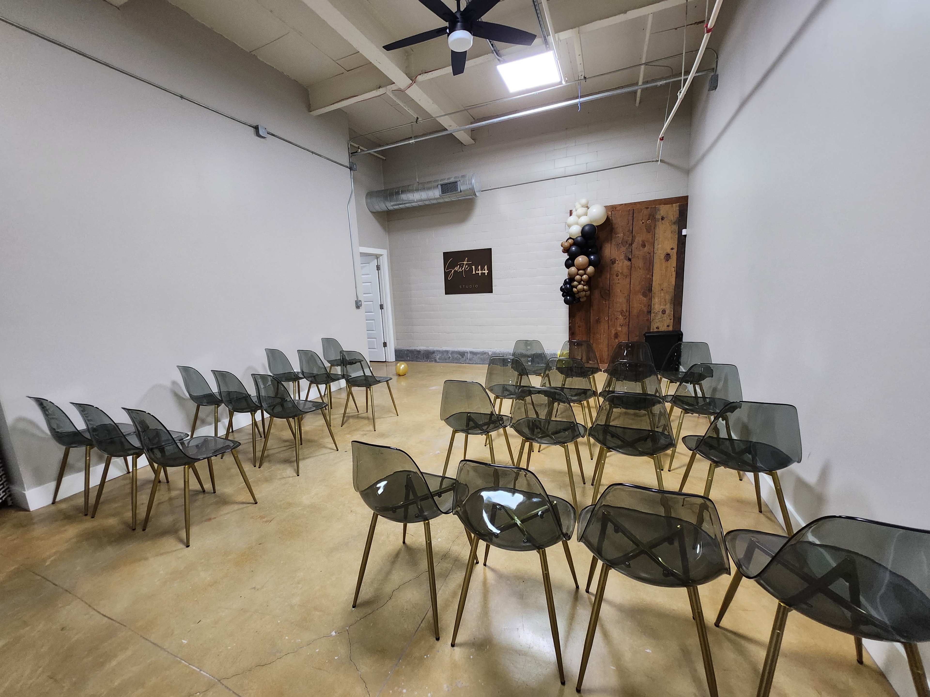 A minimalist event space with transparent chairs arranged in rows facing a wall decorated with a wood panel and a balloon arrangement.