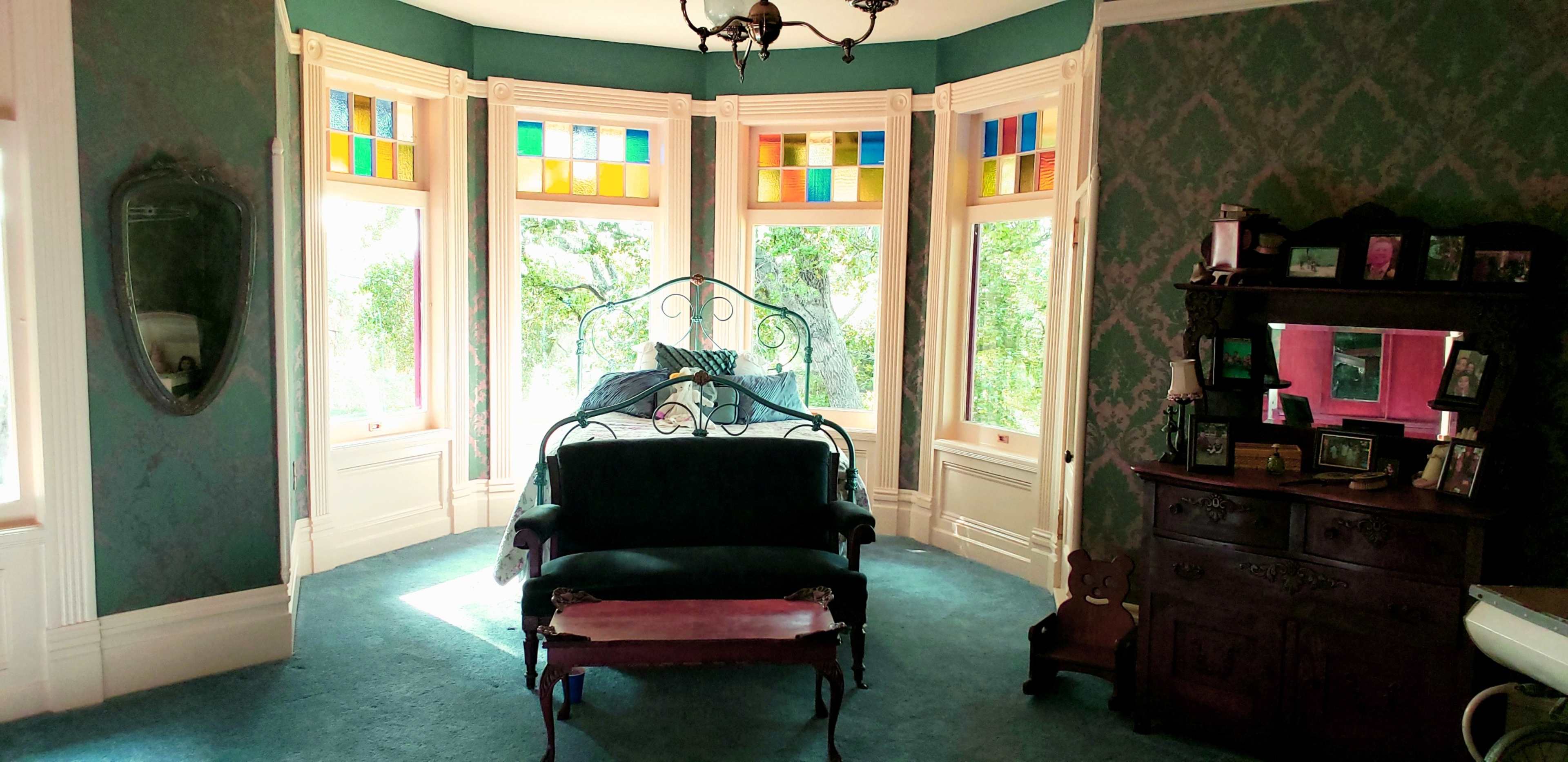 A vintage room with bay windows featuring stained glass, a wrought iron bed, a dark green wall, and antique furniture.