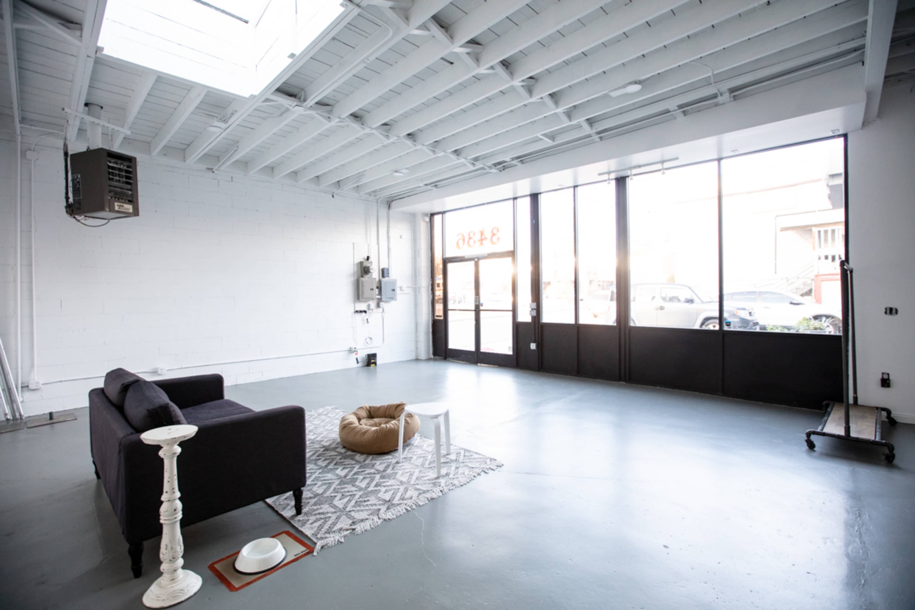 An open, minimalist interior space featuring a black sofa, a patterned rug, a small table with a lamp, and a pet bed, with large glass windows letting in natural light.