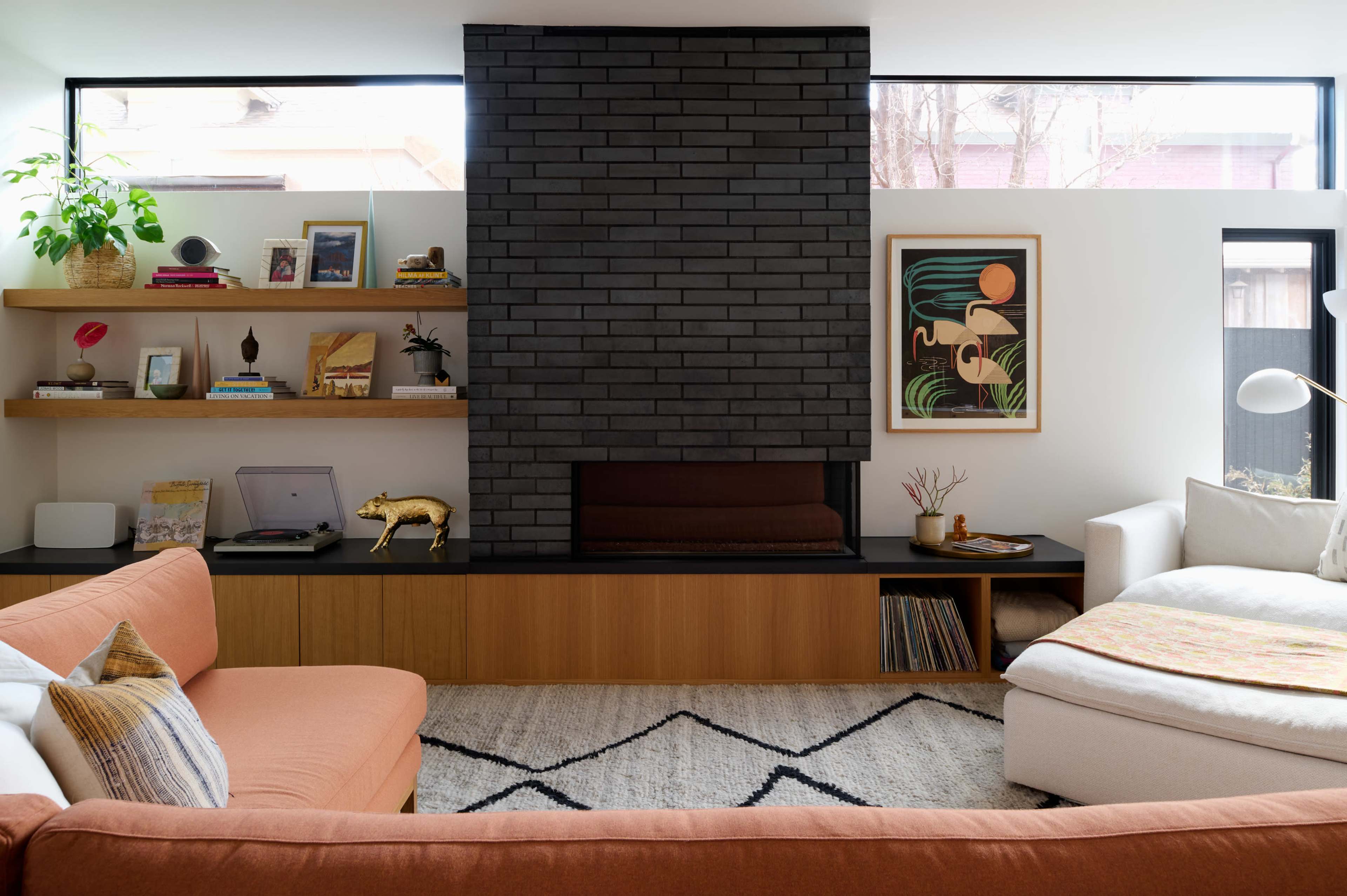 The image shows a modern living room featuring a dark brick fireplace, wooden shelves with decorative items, and a light-colored sofa paired with an orange sectional couch.