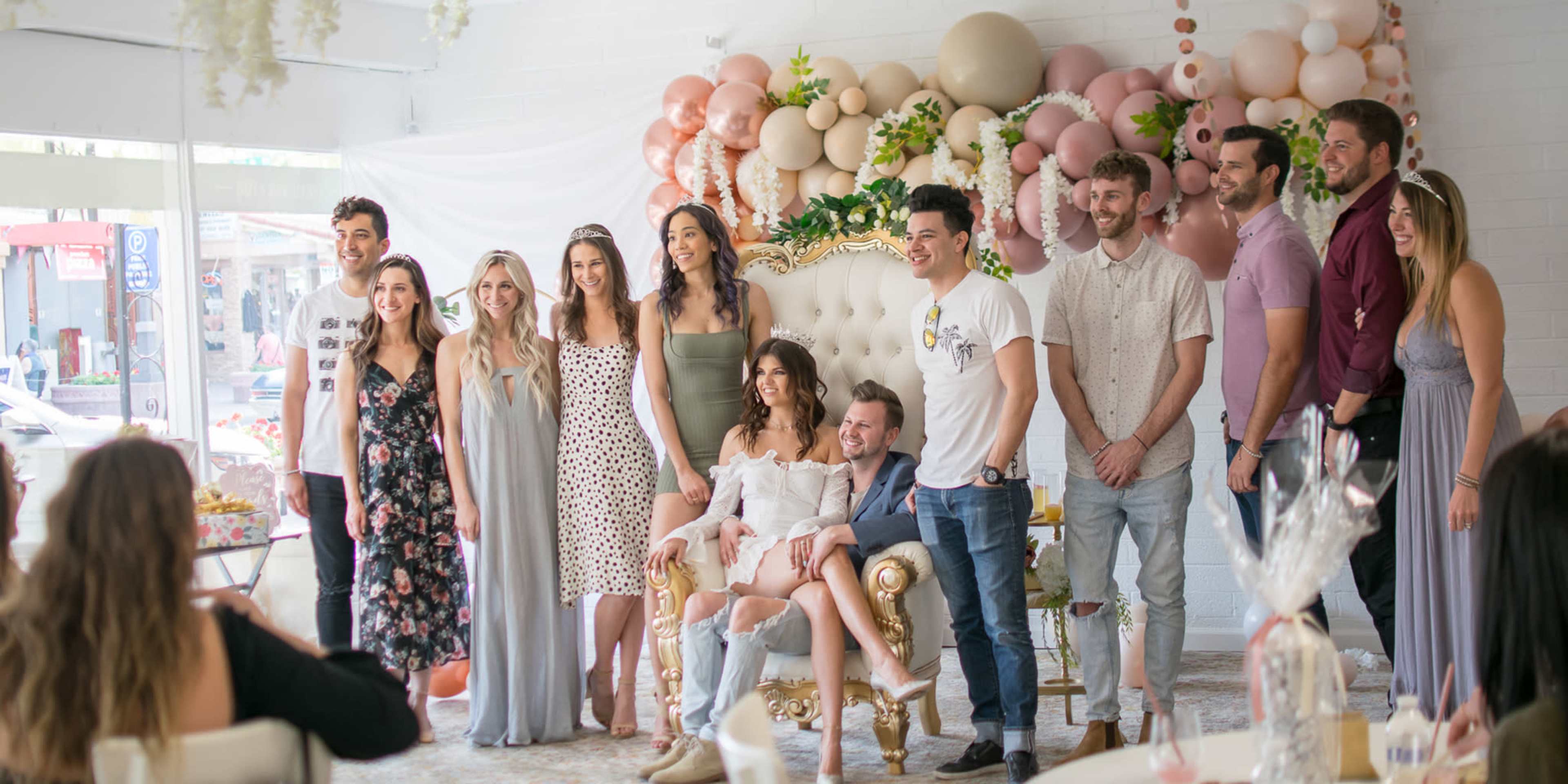 A group of people poses together around a seated woman in a decorated indoor space with balloons and floral arrangements.