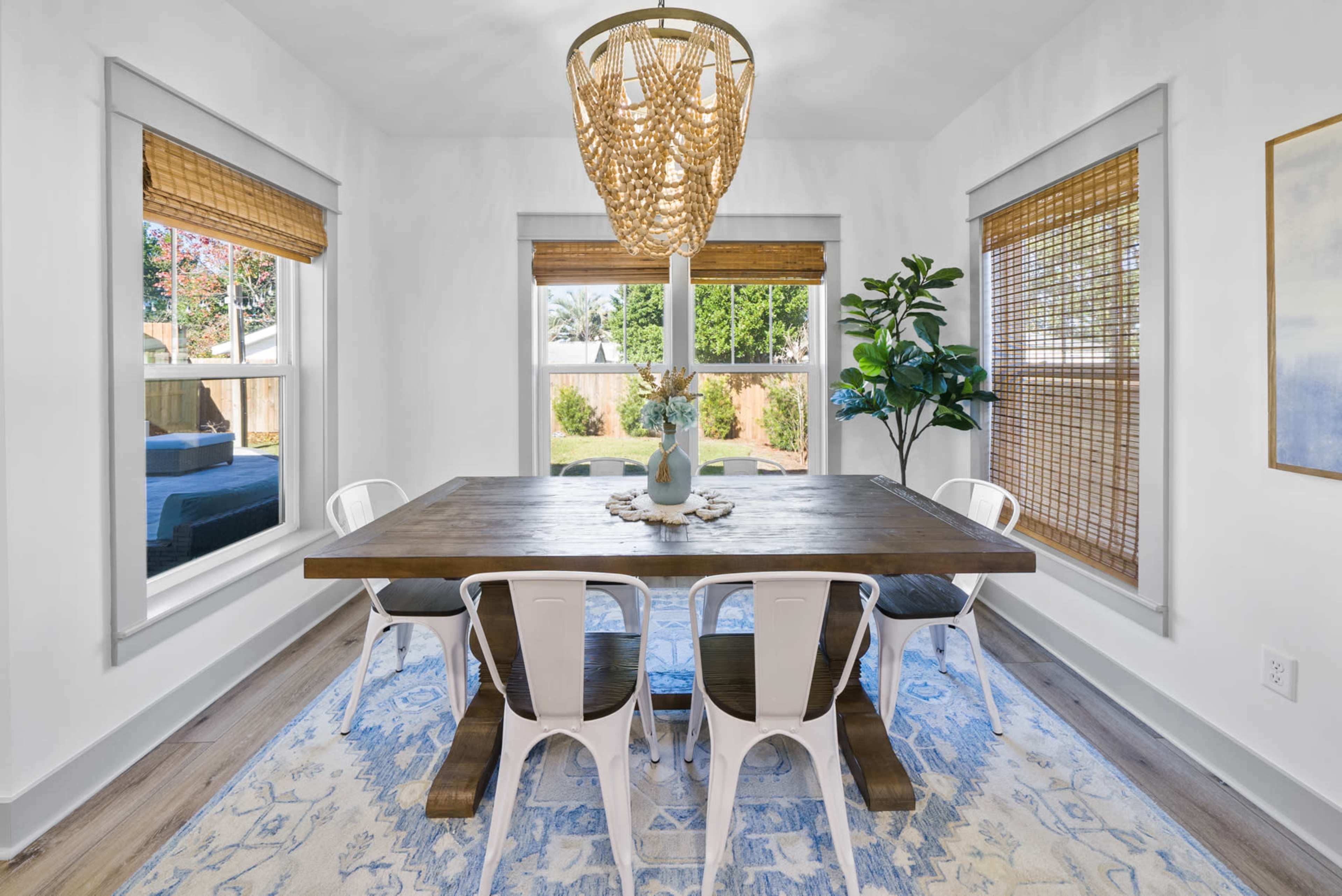 A dining room features a wooden table surrounded by white chairs, with large windows draped in bamboo shades and a modern light fixture overhead.