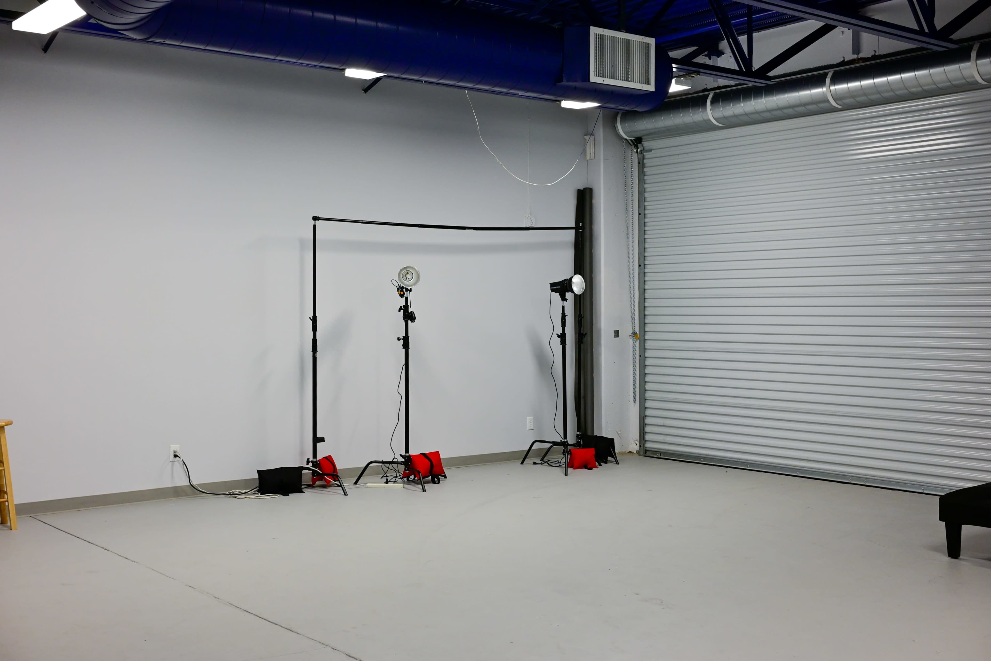 The image shows a spacious, empty room with a gray floor and walls, featuring two lighting stands and a backdrop frame near a large closed garage door.