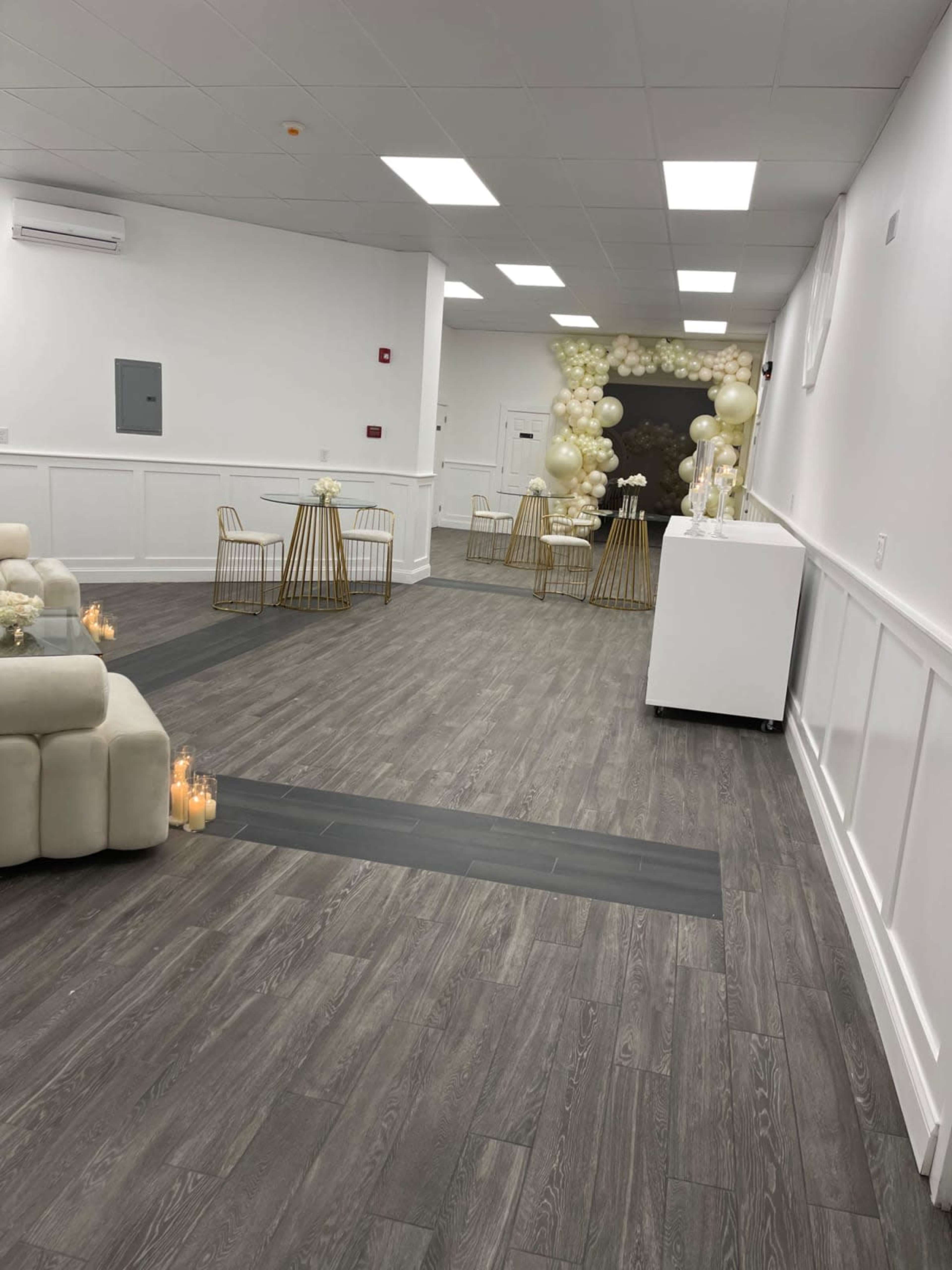A spacious, well-lit event room featuring a modern seating area, gold tables, and a decorated backdrop with balloons.
