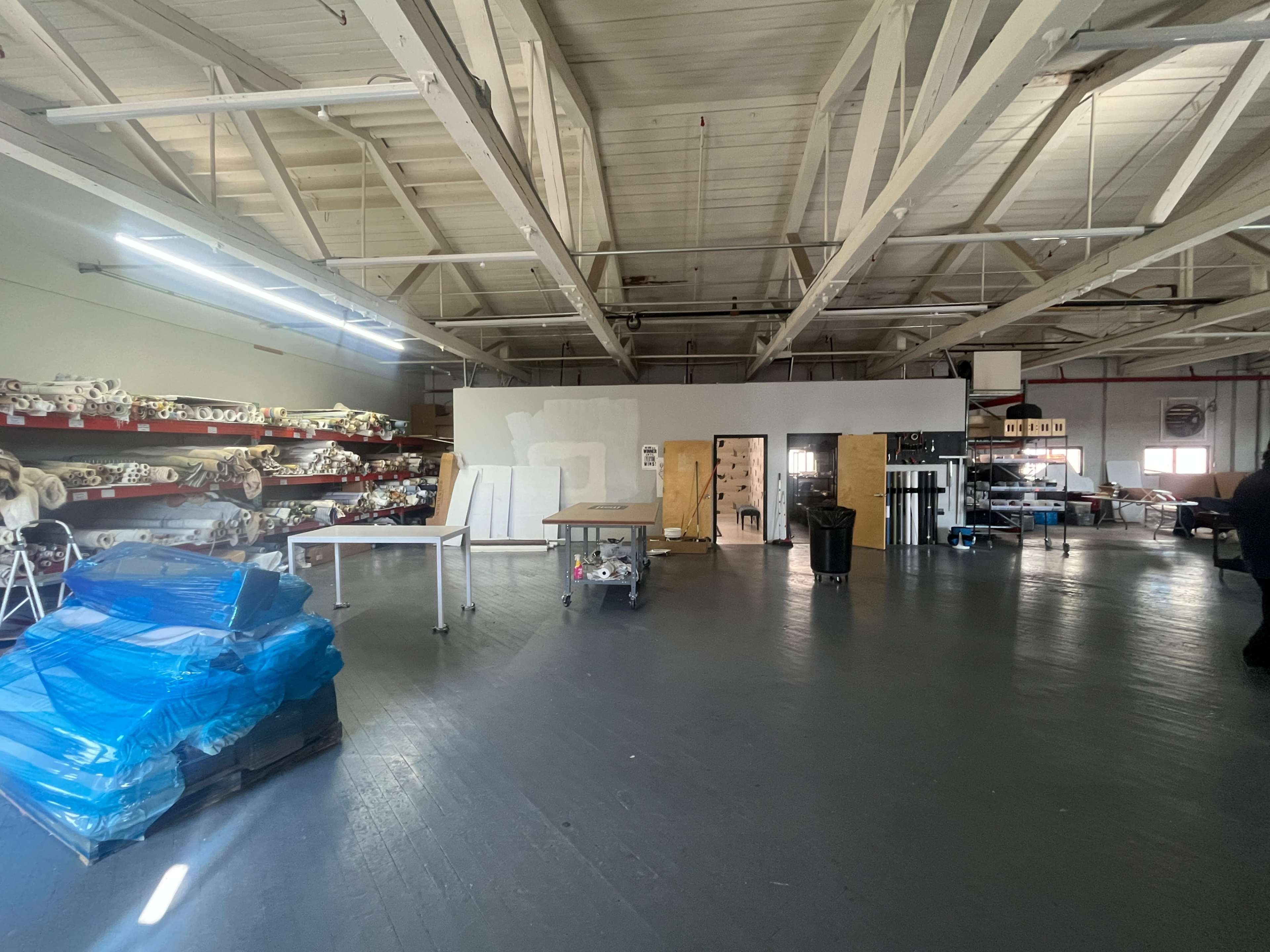 The image shows a spacious industrial interior with high ceilings, featuring shelves stocked with rolls of material and various work tables scattered throughout.