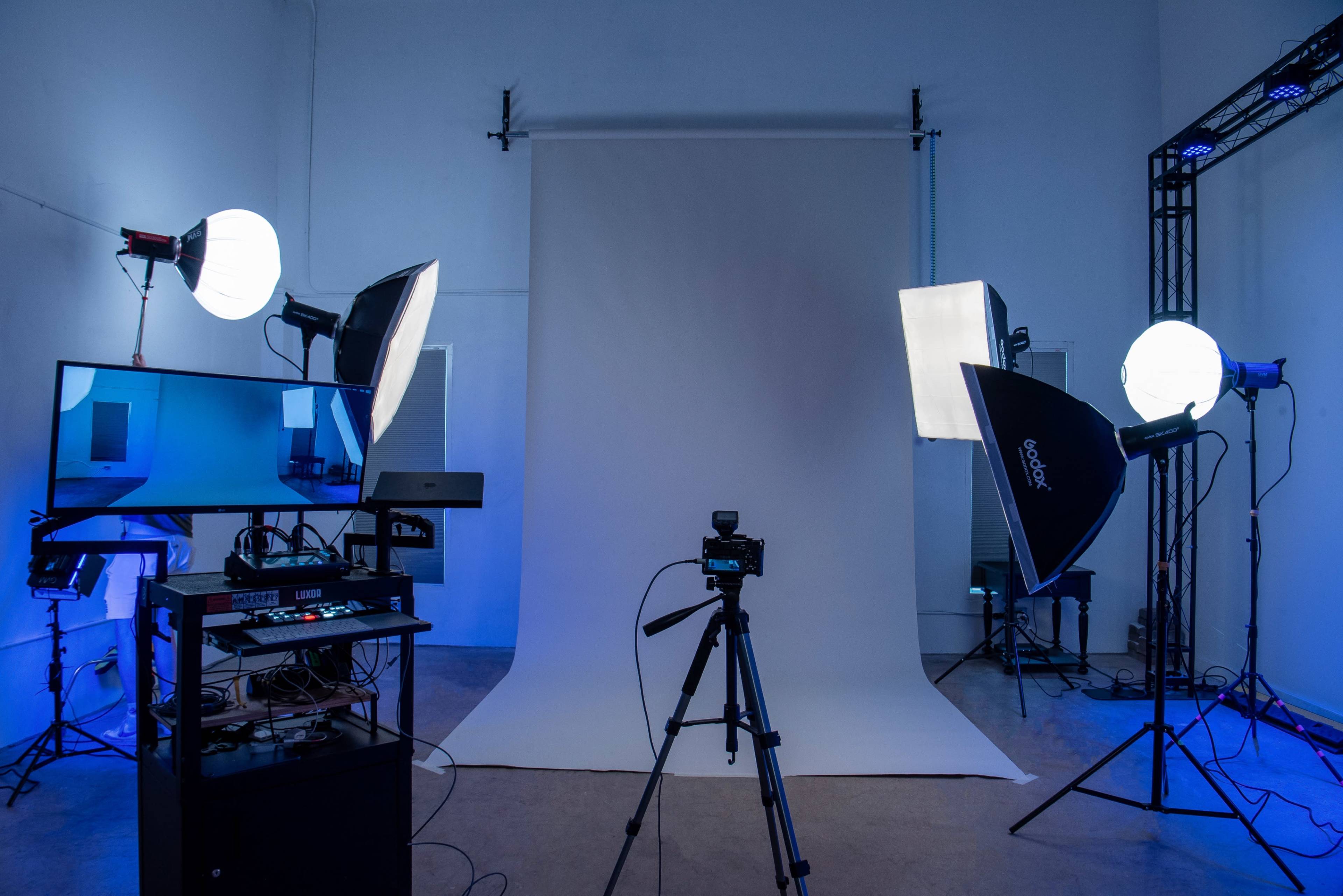A photography studio setup features a white backdrop, various lighting equipment, a camera on a tripod, and a computer monitor.