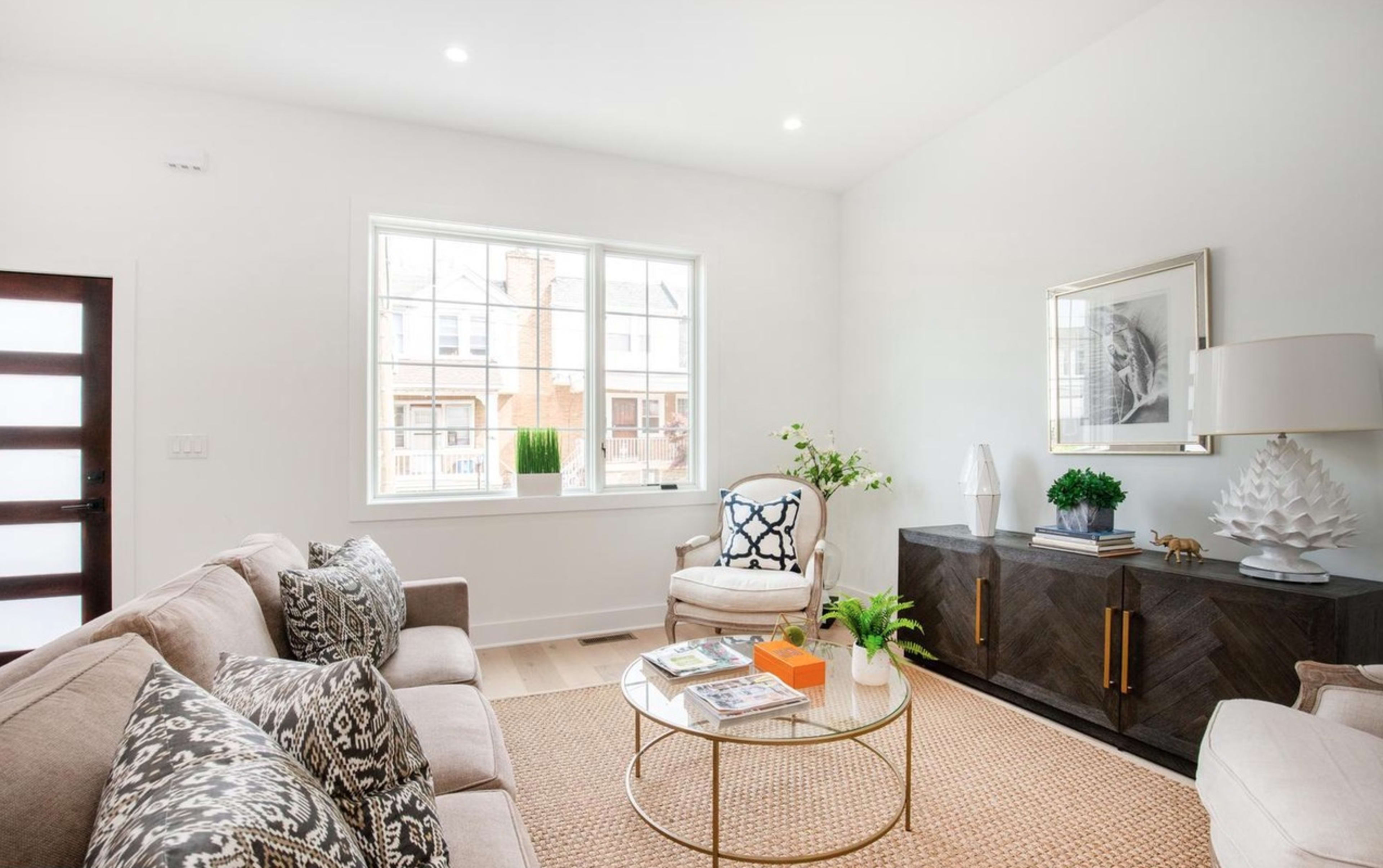 The image shows a bright and modern living room featuring a beige sofa, a decorative rug, a round coffee table with magazines, and a dark wooden sideboard under a large window.