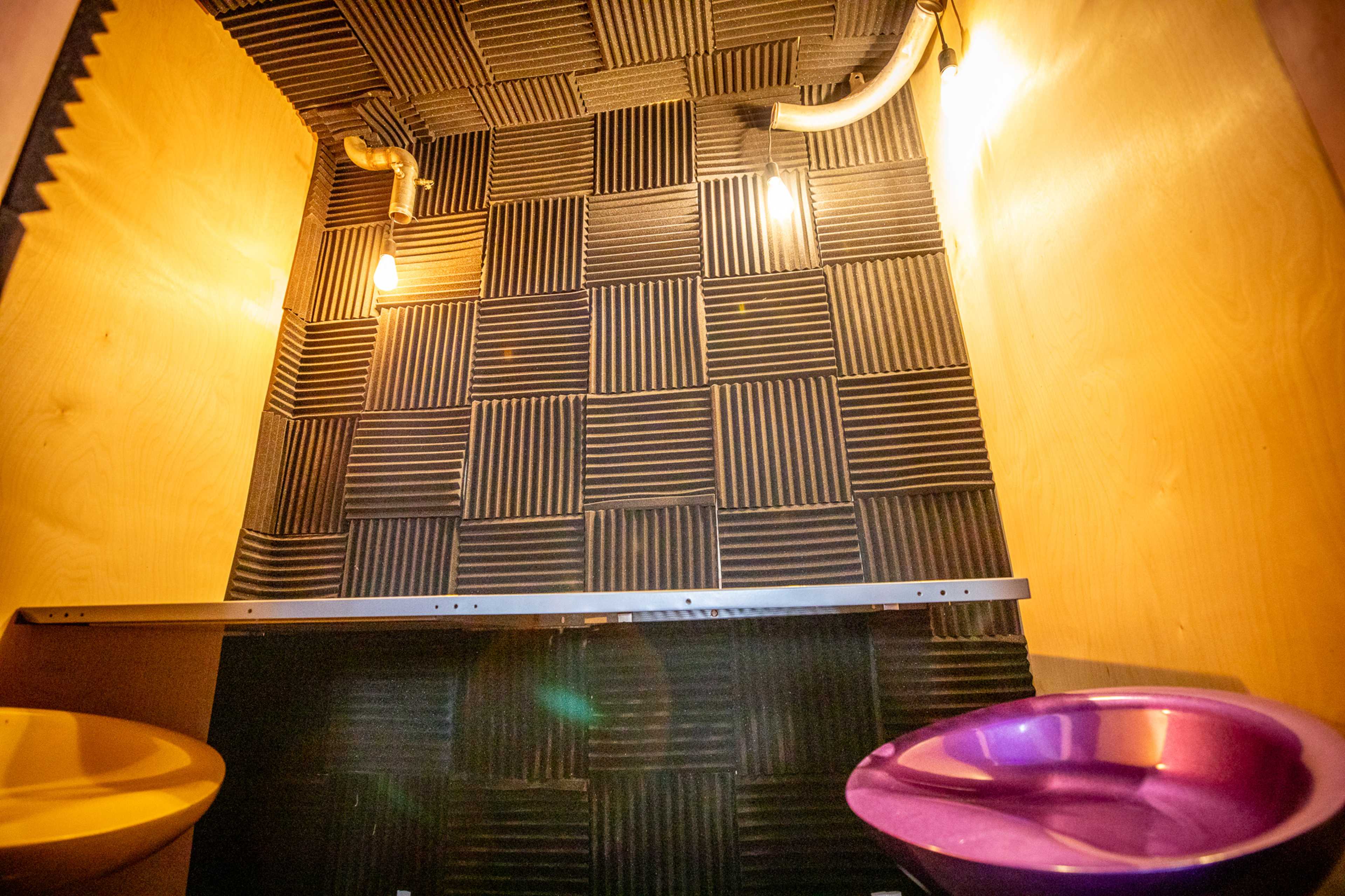 The image shows a small room with black acoustic foam tiles on the walls, a wooden bench, and two differently colored toilets.