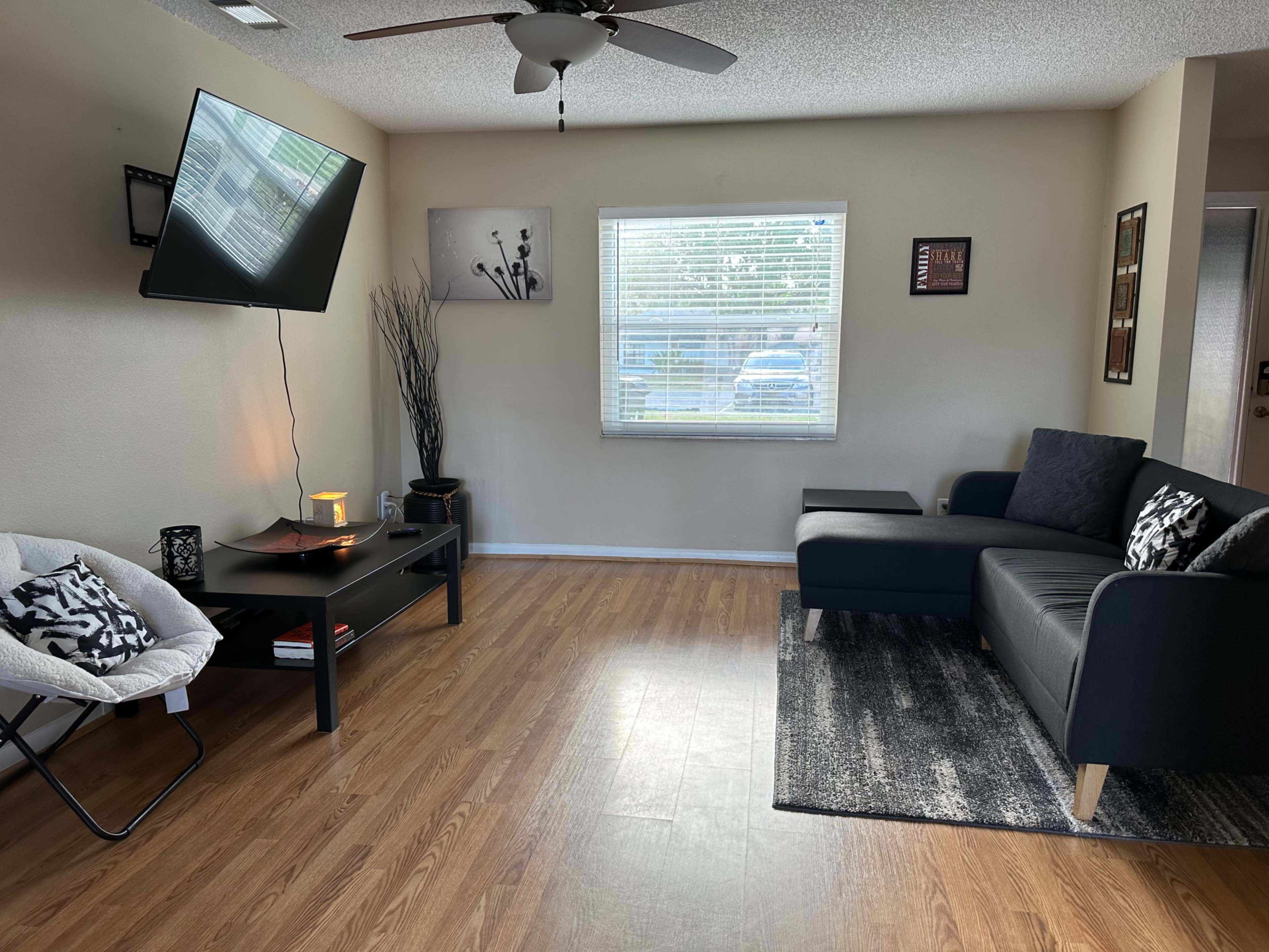 The image shows a living room with a black sectional sofa, a coffee table, a floor lamp, and a wall-mounted television, all arranged on a wooden floor under a ceiling fan.