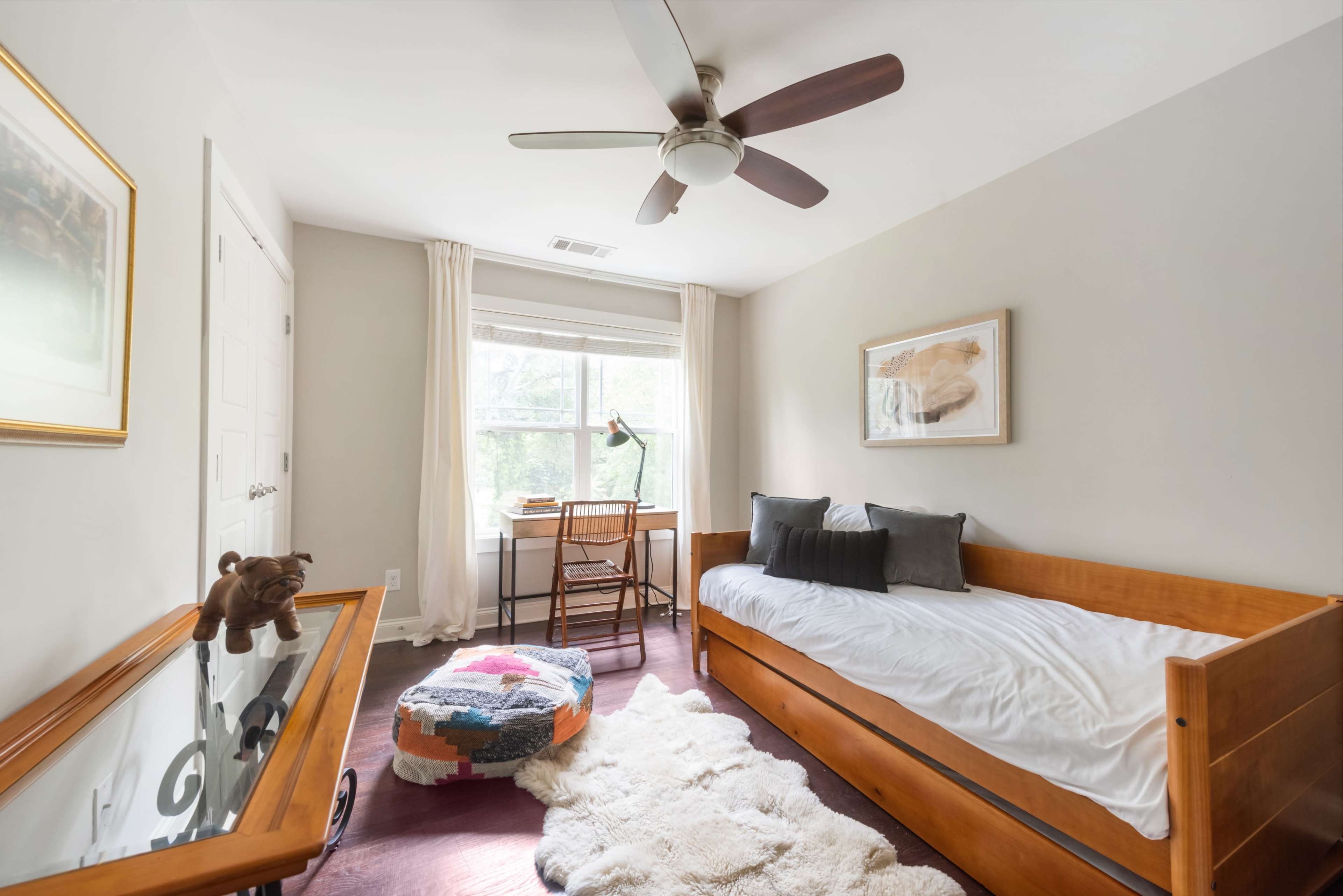 The image shows a simple bedroom featuring a wooden daybed, a small desk by the window, a colorful pouf, and soft lighting from a ceiling fan.