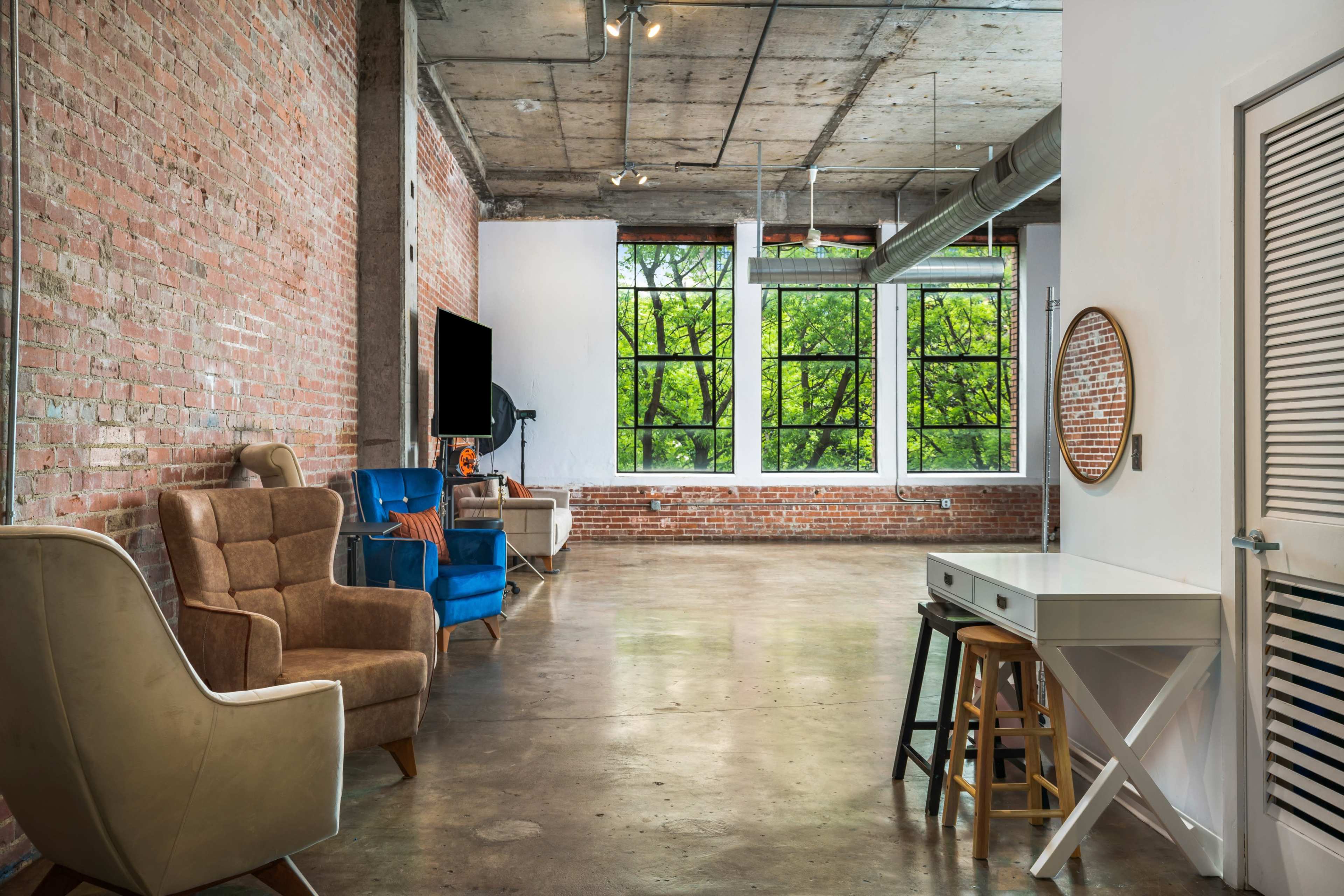 A spacious room features exposed brick walls, large windows overlooking greenery, and a mix of seating options including an armchair and a blue chair.