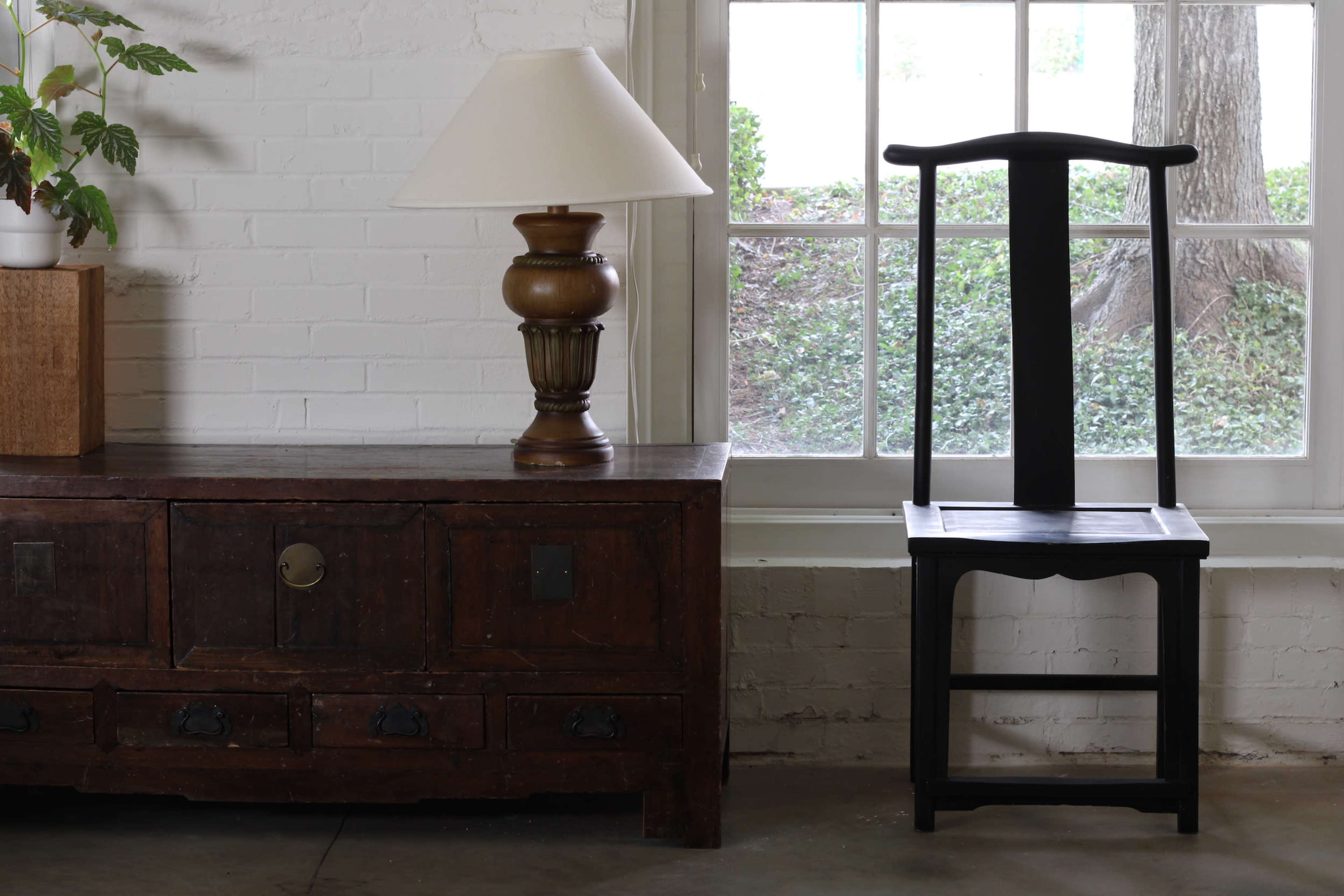 A wooden side table with a lamp stands beside a black chair against a white brick wall, illuminated by natural light coming through a window.