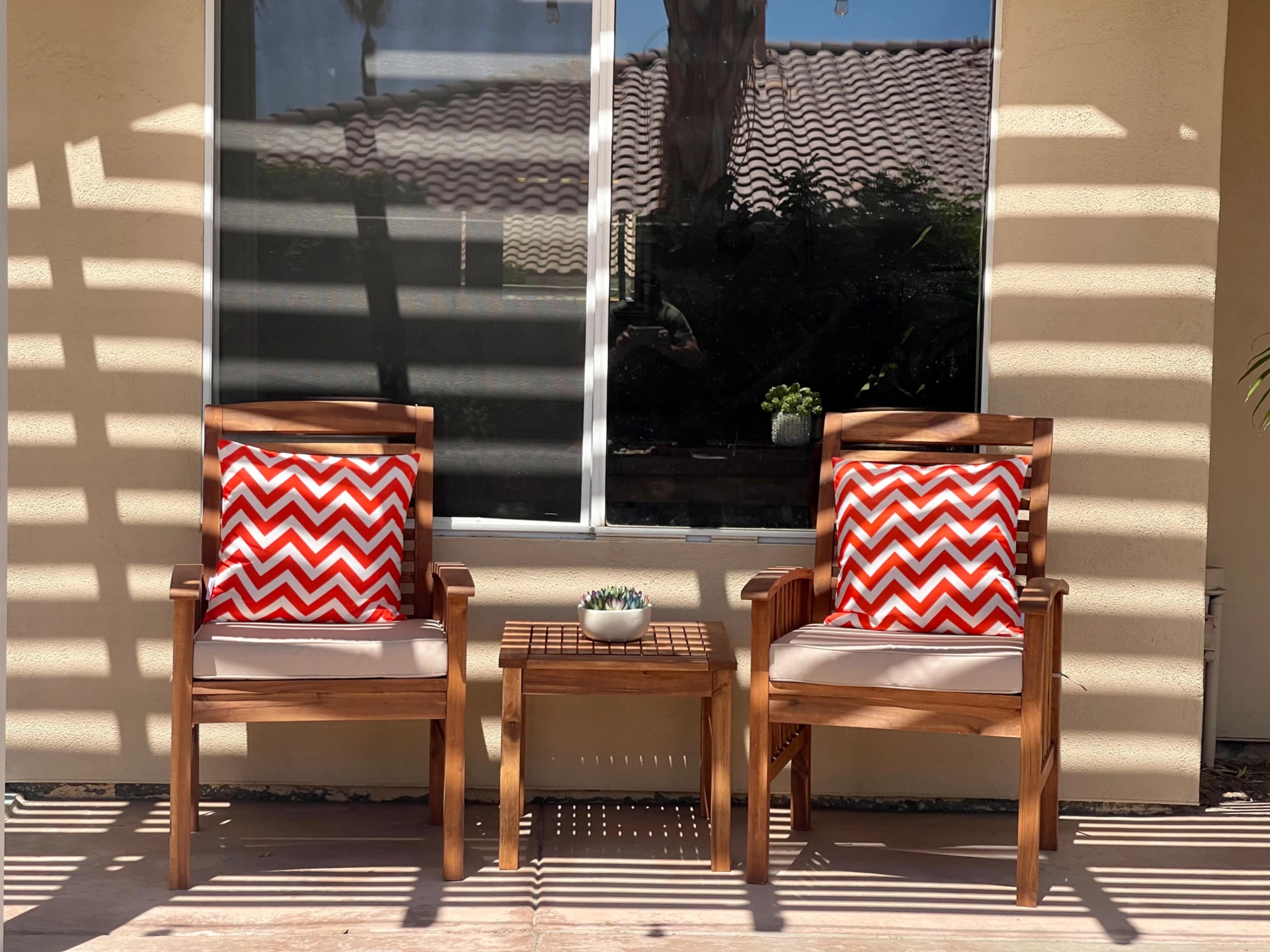 Two wooden chairs with red and white chevron-patterned cushions are positioned beside a small table in front of a window, with sunlight casting striped shadows across the scene.